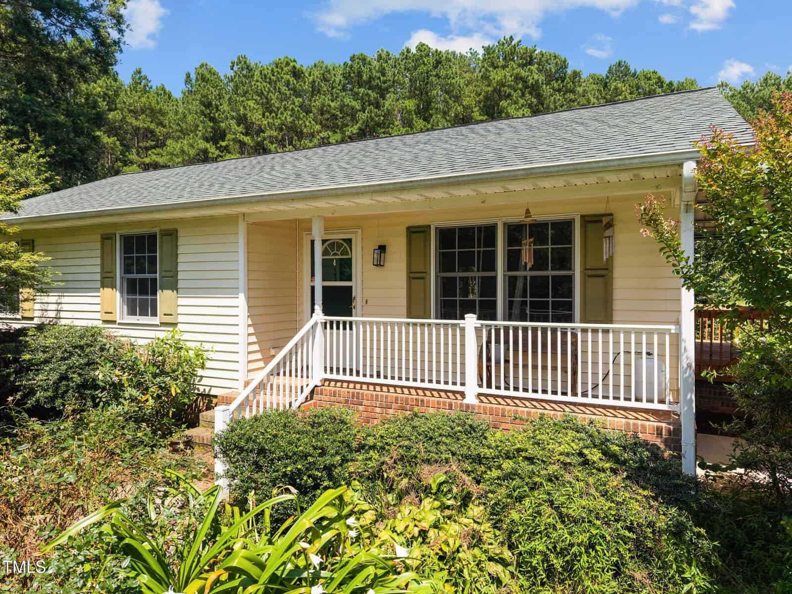 A one-story house with light yellow siding, green shutters, and a covered front porch with white railings, surrounded by lush greenery and shrubs, set against a backdrop of tall trees.