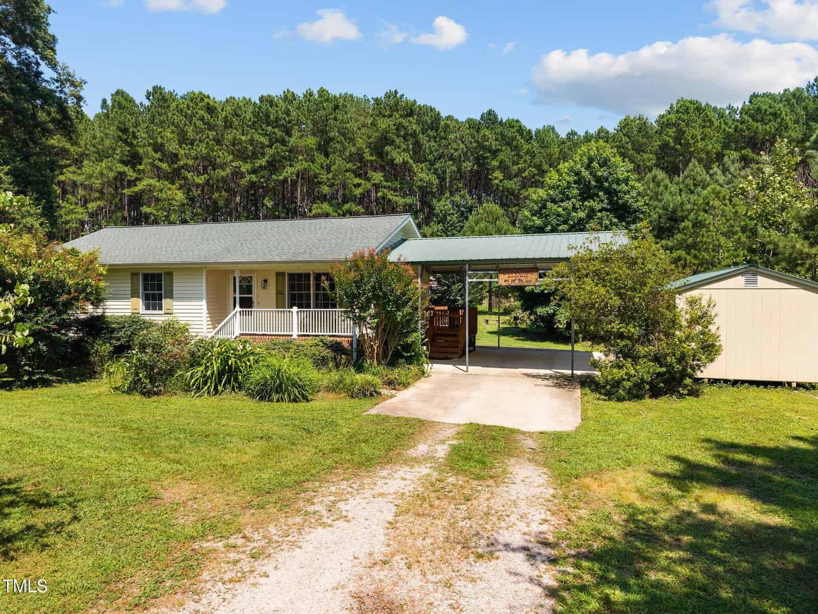 Single-story house with a front porch, carport, and large shed, surrounded by green lawn, shrubs, and trees, with a dense forest in the background under a blue sky with clouds.