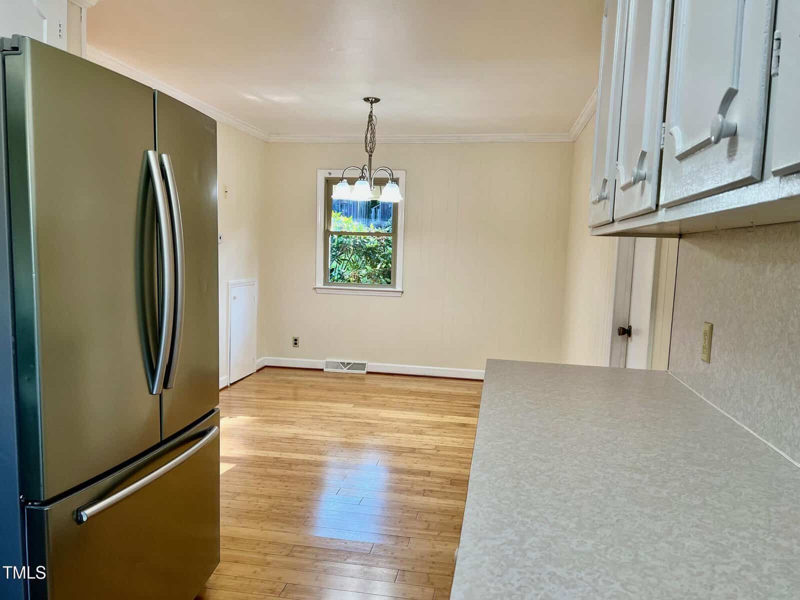 Bright kitchen with wooden floors, white cabinets, stainless steel fridge, and a small dining area under a three-light chandelier next to a window with green foliage visible outside.