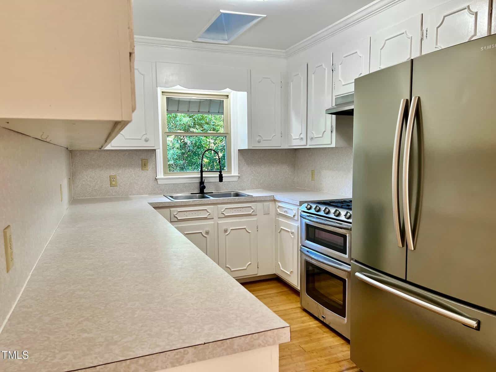 Bright kitchen with white cabinets, stainless steel fridge and oven, double sink below a window, light countertops, and wood flooring. The space looks clean and modern with natural light from the window.