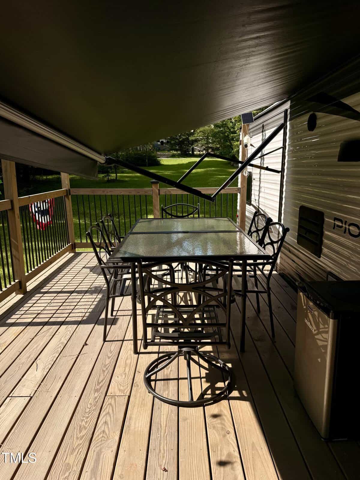 A glass-top table with six metal chairs sits on a wooden deck next to a camper. An awning provides shade, and a green lawn with trees is visible beyond the deck railing.
