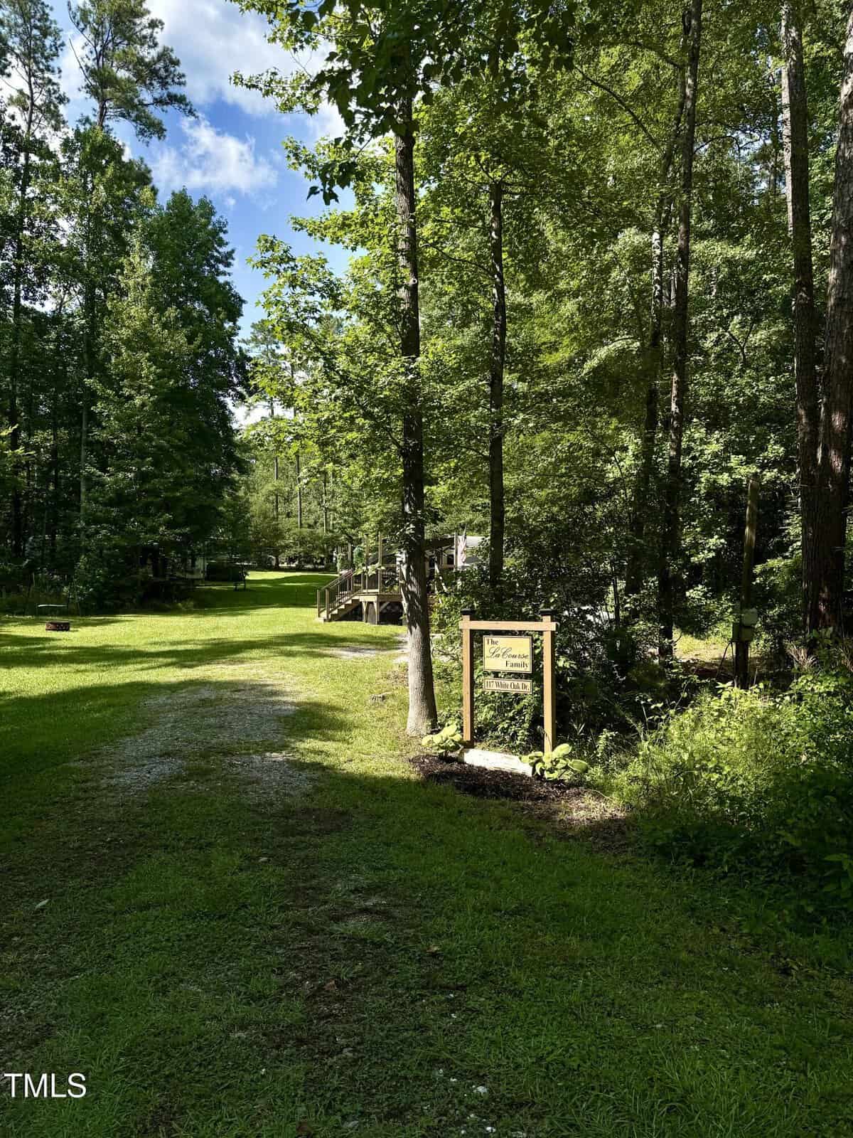 A sunlit, grassy path leads through a wooded area with tall trees. A wooden sign with illegible text stands by the path, and a wooden bridge is visible in the background among the greenery.
