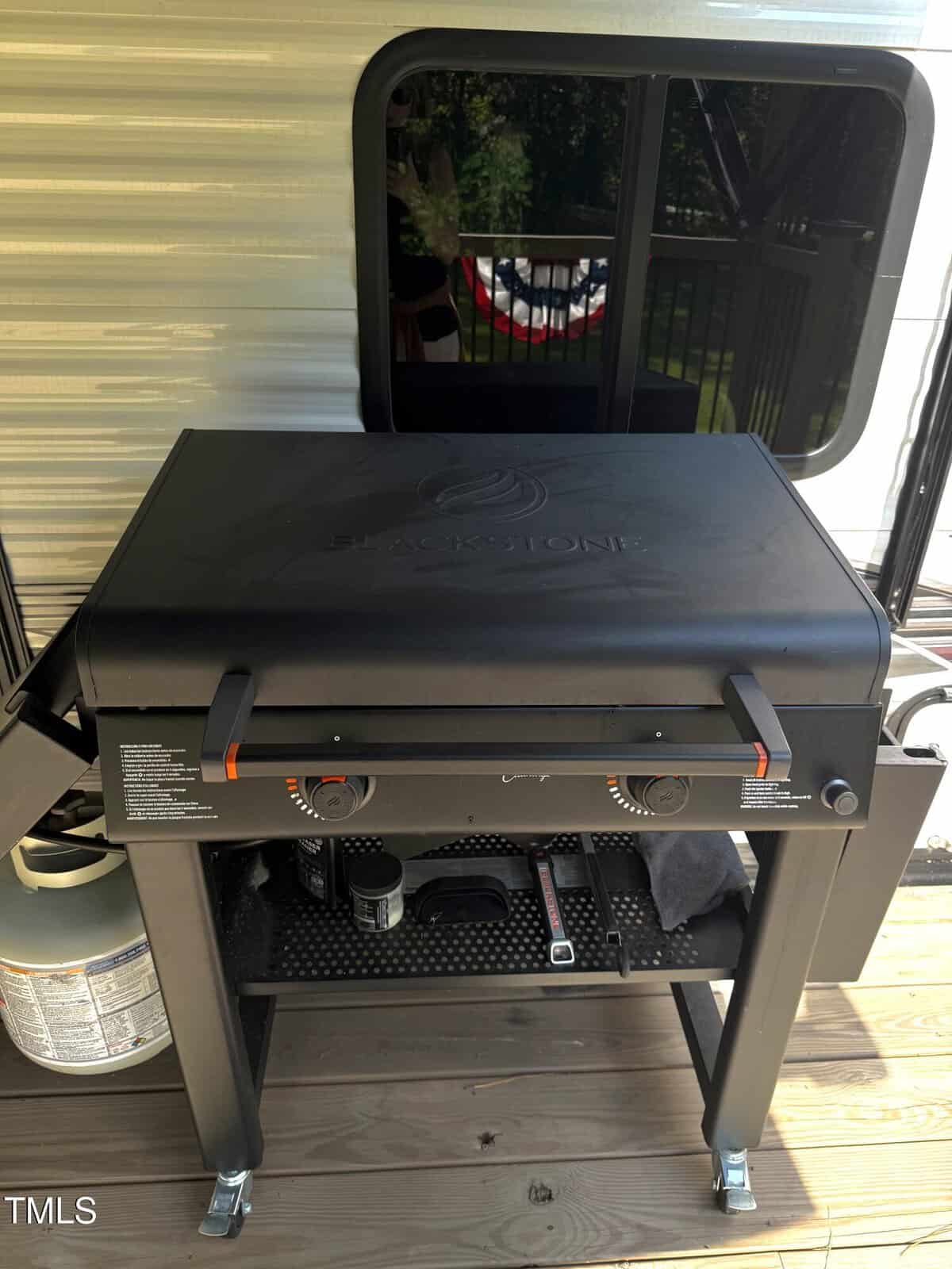 A Blackstone outdoor gas griddle with a closed lid sits on a wooden deck next to a trailer. A propane tank is attached on the left side, and various grilling tools are stored on the shelf below.
