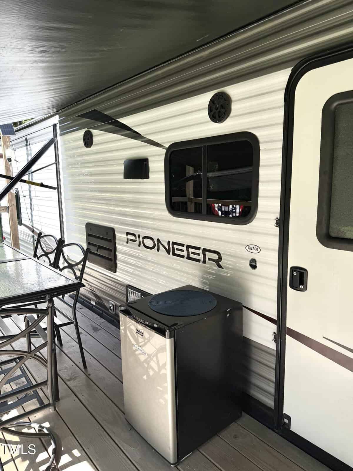 Side view of a Pioneer RV with an open window, glass table with chairs, and a small stainless steel mini fridge placed outside on a wooden deck under an awning.