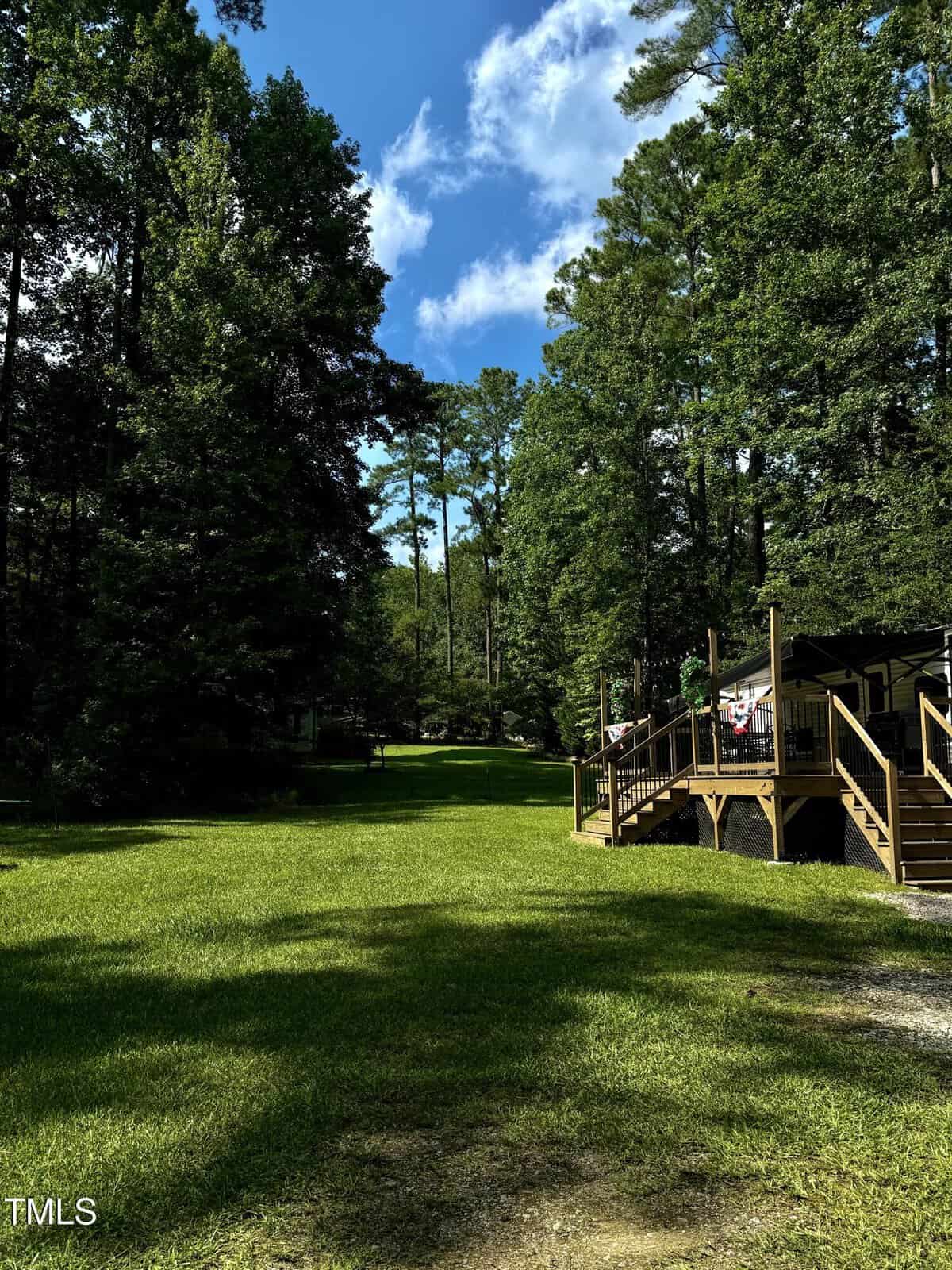 A lush green lawn bordered by tall trees under a blue sky with scattered clouds; a wooden deck with railings and American flags is visible on the right side of the image.