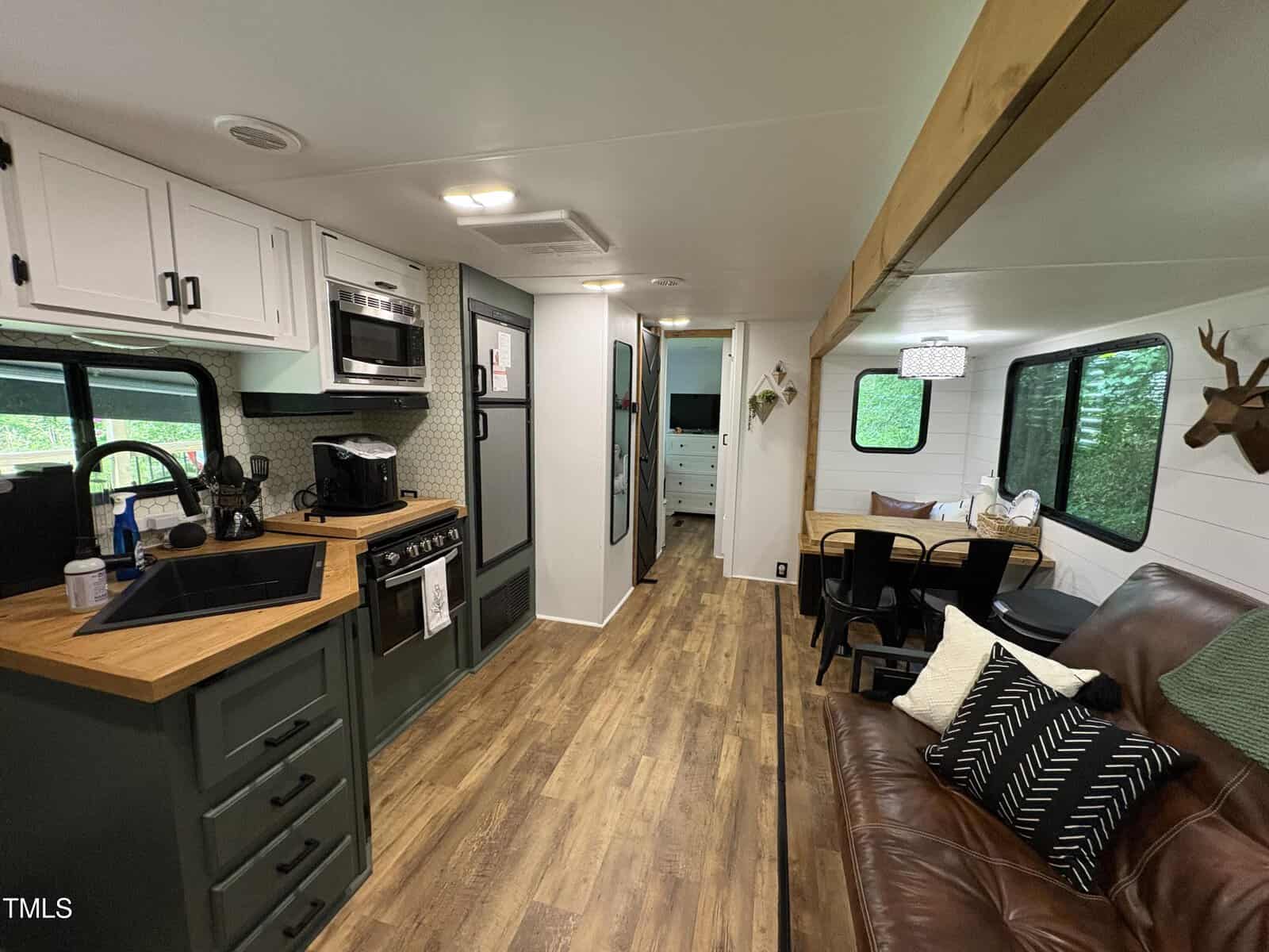 Interior of a renovated RV with wood floors, green lower cabinets, white upper cabinets, a kitchen area, dining nook, brown leather sofa, and windows with outdoor views. Decor includes pillows and a faux deer head.