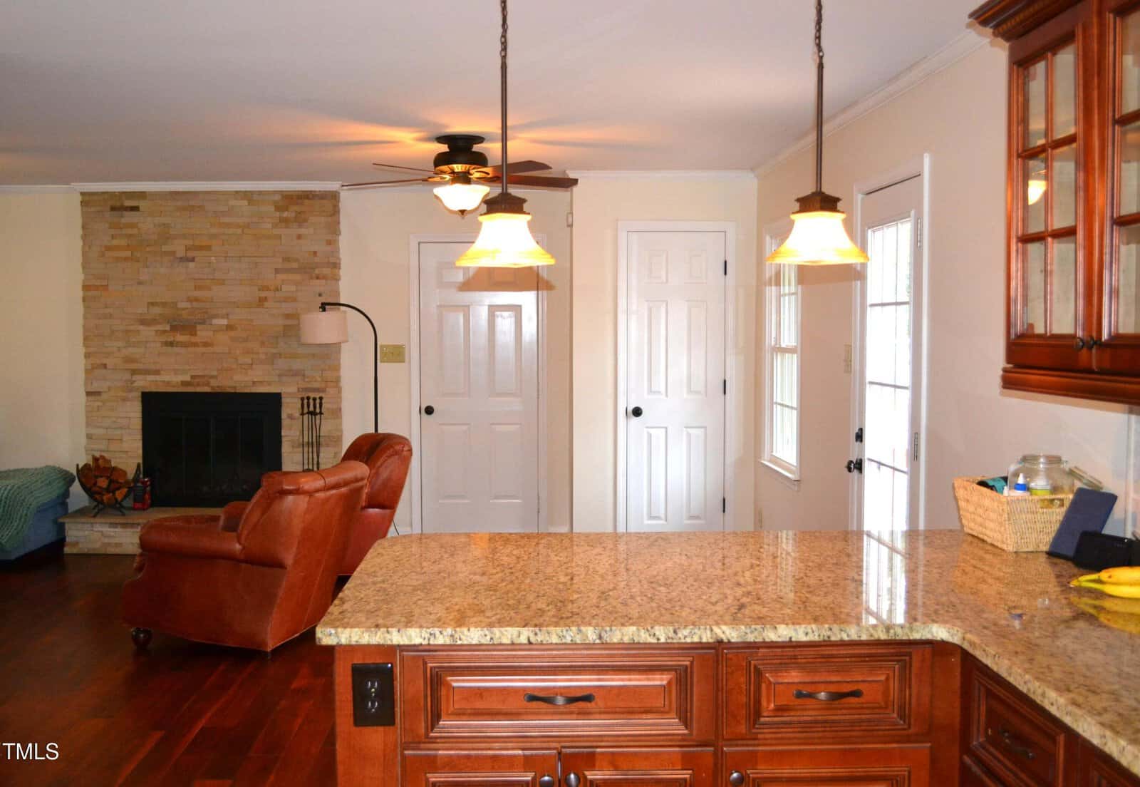 A view from a kitchen with granite countertops and wooden cabinets, looking into a living room with a stone fireplace, brown leather recliner, ceiling fan, and two white doors on the back wall.