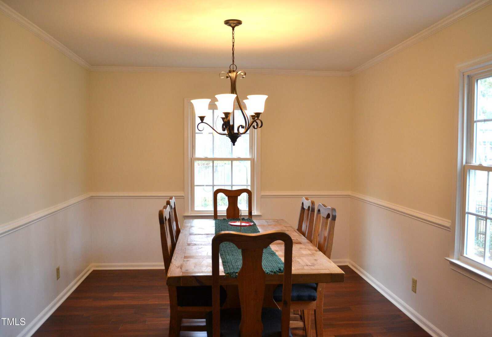 A dining room with cream-colored walls, a wooden table, six chairs, a chandelier above, and sunlight streaming through two windows in the background.