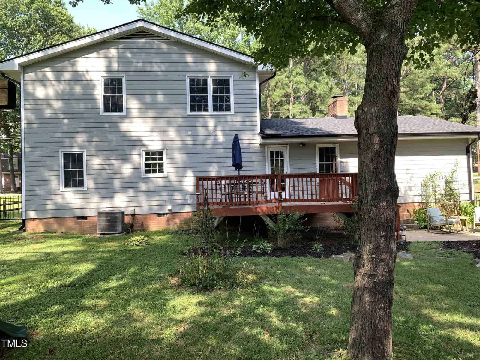 A two-story house with light gray siding, a brick foundation, multiple windows, and a wooden deck with a blue umbrella in the backyard. There is a tree and green lawn in the foreground.