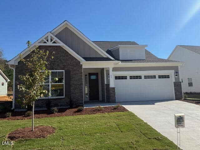 A modern single-story house with stone and siding exterior, attached two-car garage, and a small tree in the front yard sits at 35 Sawtooth Oak Lane, Youngsville. A “For Sale” sign is displayed in the window, while bright sunshine and a clear blue sky provide a welcoming backdrop.