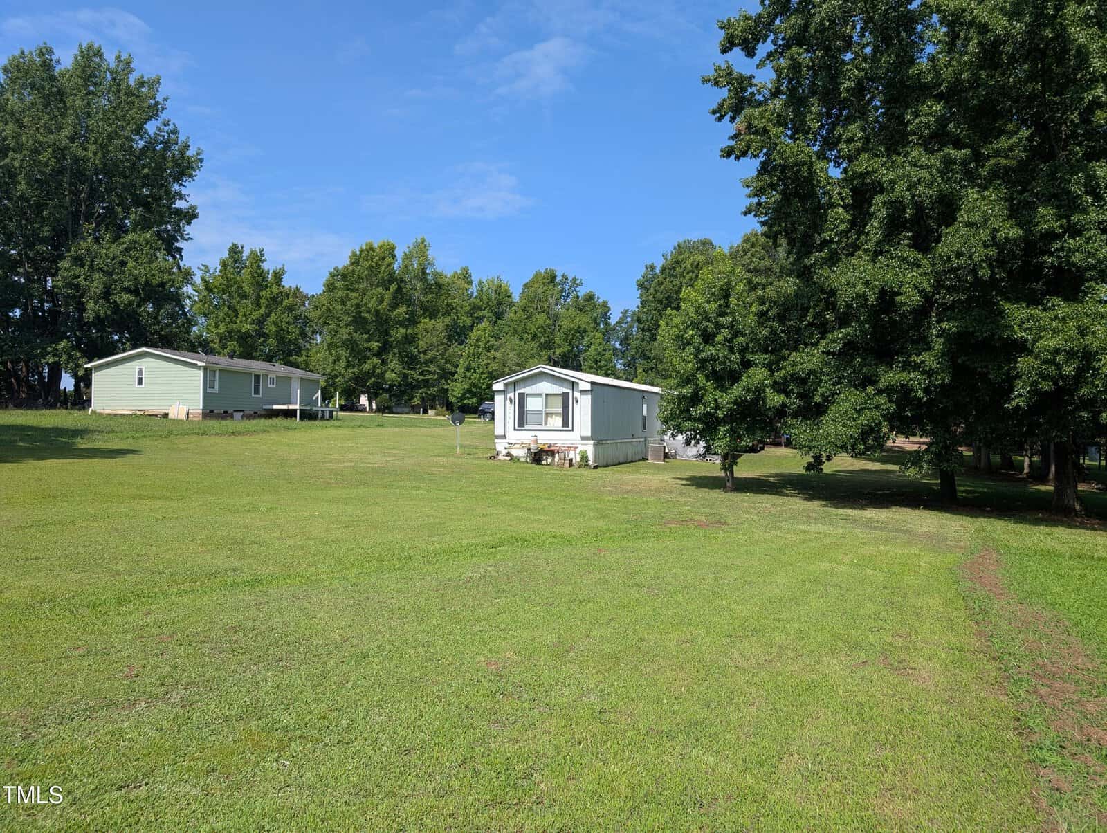 A single-story manufactured home sits on a large grassy lawn with trees in the background and another similar home to the left; the sky is clear and blue.