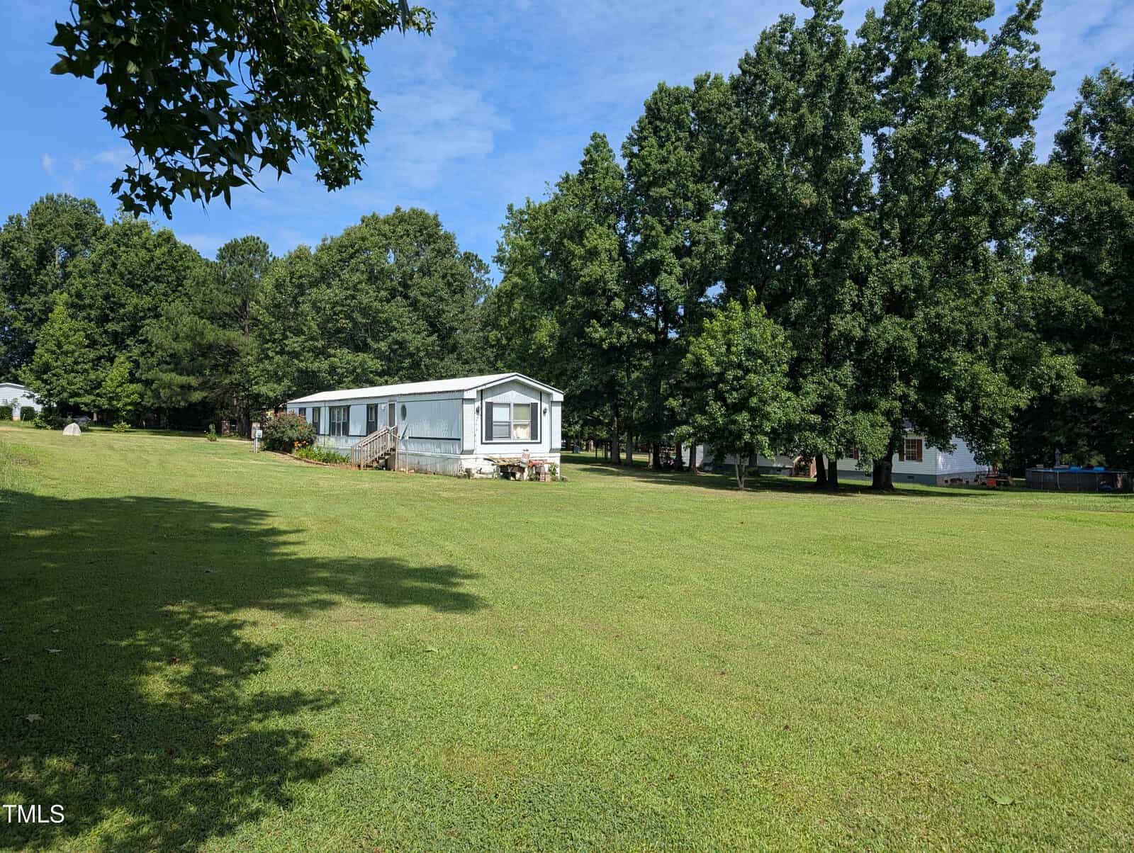 A light blue manufactured home with white trim sits on a large, grassy lot surrounded by mature trees under a blue sky. A small wooden porch leads to the front door.