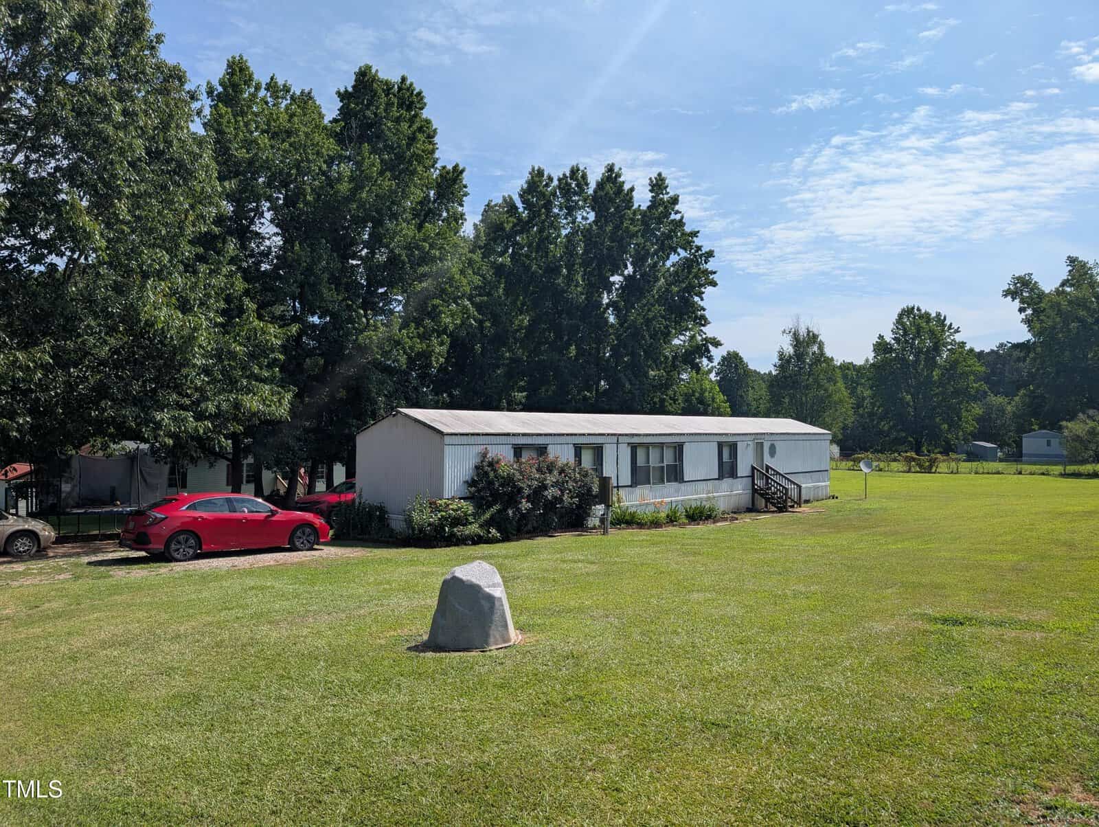 A white single-wide mobile home sits on a large grassy lot at 1506 Pocomoke #2 Drive, Franklinton, with a red car parked to the left. Trees line the background and a large rock rests on the lawn under a mostly clear sky with some clouds.