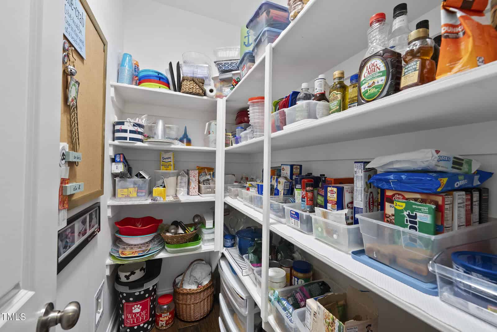 A neatly organized pantry with white shelves holding various food items, spices, condiments, and kitchen containers. Small kitchen appliances and baskets are on the lower shelves; colorful dishes and bins are visible on the upper shelves.