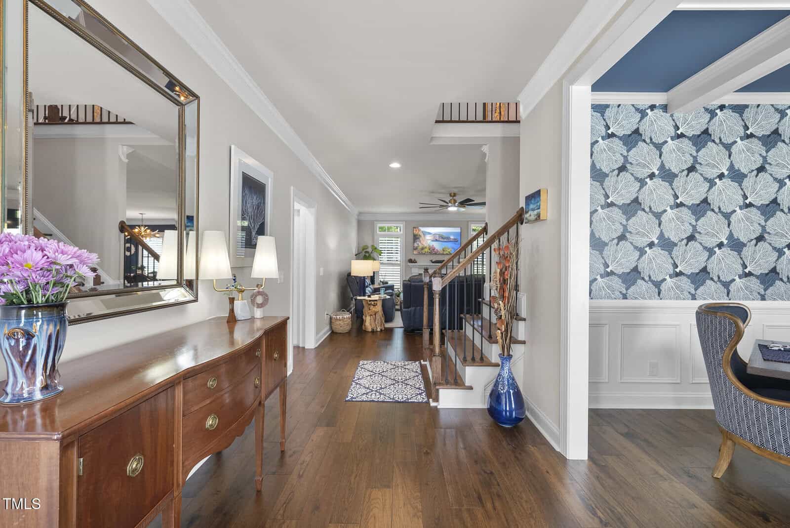 A hallway with hardwood floors, wooden console table, mirror, lamps, and flower vase. A staircase with wooden railing is on the right, leading up. Adjacent room has blue floral wallpaper and white wainscoting.