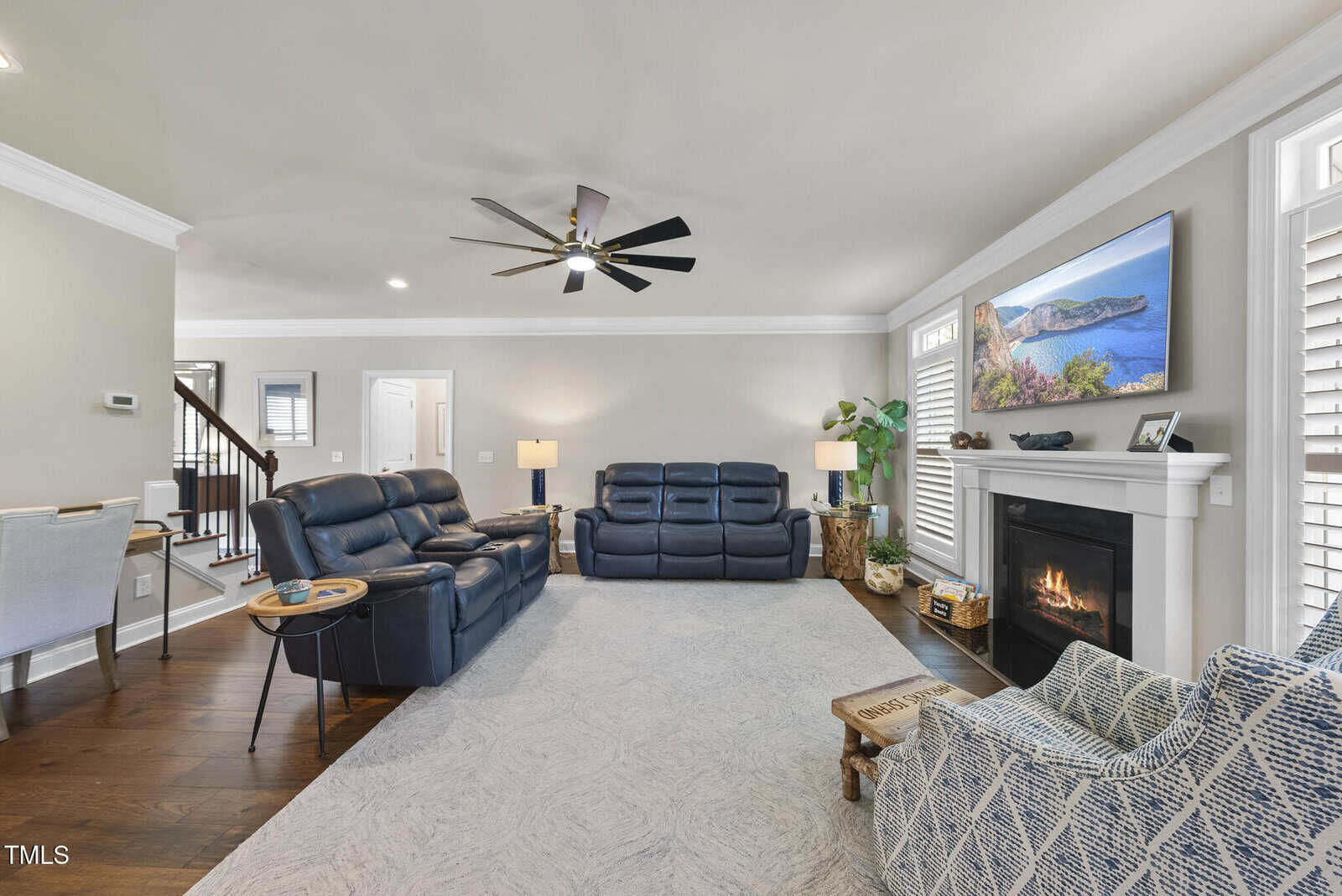 A modern living room with dark wood floors, two blue leather reclining sofas, a patterned armchair, a fireplace with a TV above it, a ceiling fan, and neutral-colored walls with bright lighting.