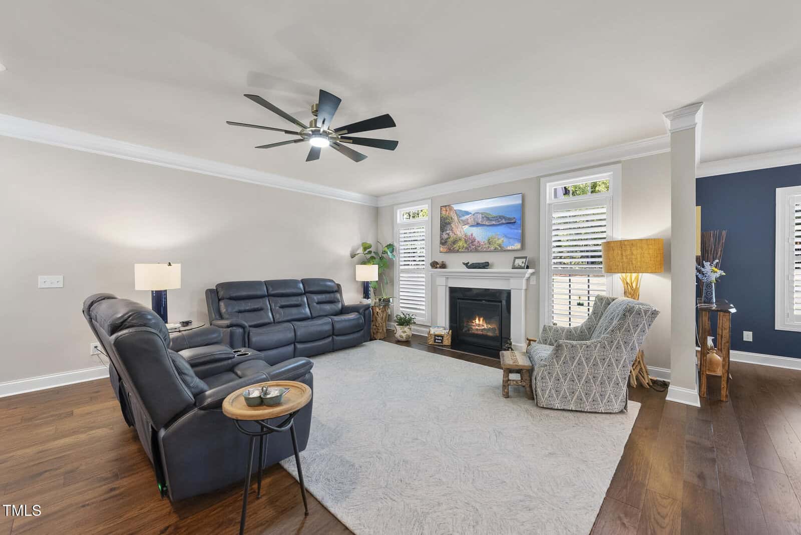 A spacious living room with two black leather sofas, a patterned armchair, a fireplace with a TV above it, wood floors, white walls, large area rug, and ceiling fan. Natural light enters through shuttered windows.