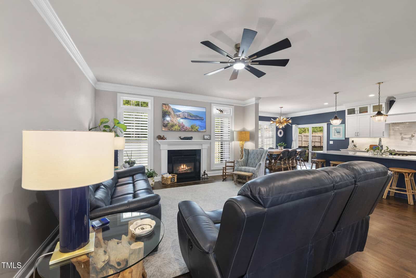 Modern living room with dark leather sofas, a glass side table, a fireplace with a painting above it, and an open view to the dining area and kitchen with bar stools and hanging lights. Ceiling fan is on.
