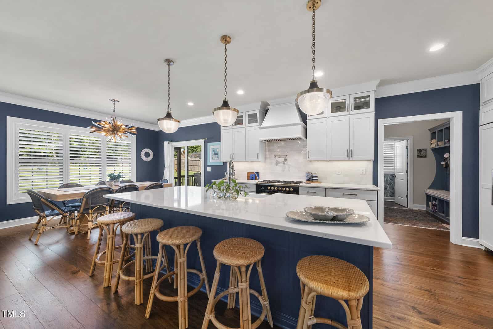 Modern kitchen with navy blue walls, white cabinets, a large white island with wicker barstools, pendant lights, hardwood floors, and a dining area with a wooden table and unique chandelier.