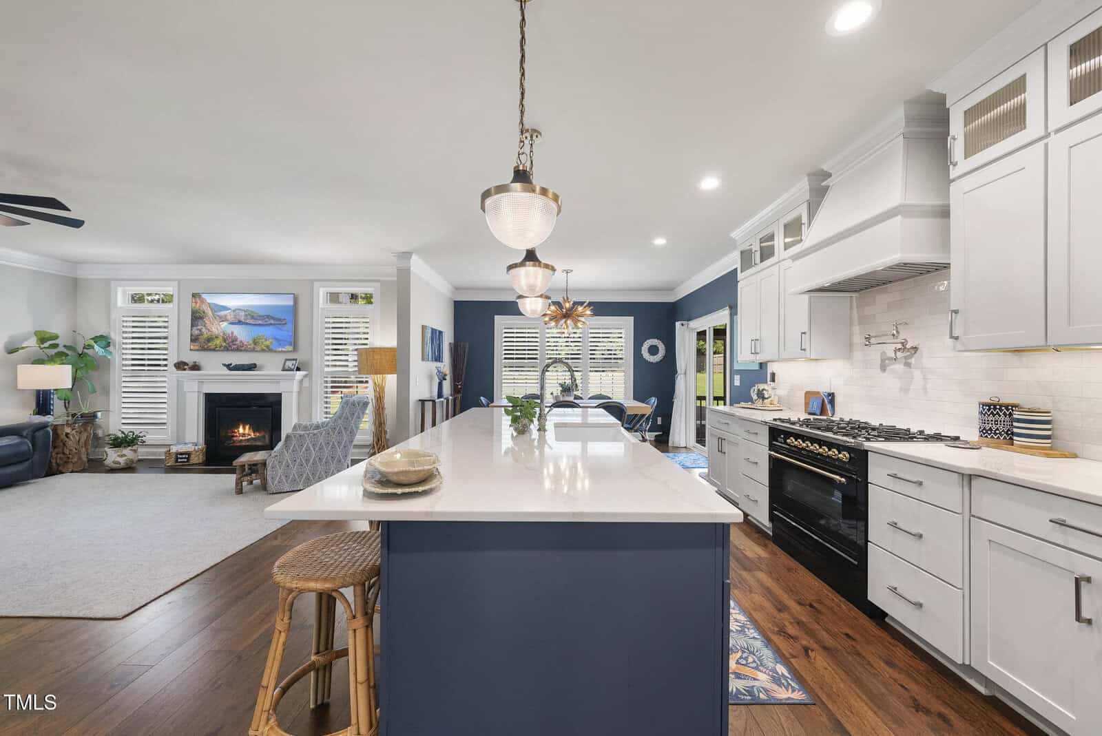 A modern open-concept kitchen with a large island, white cabinets, stainless steel appliances, and hardwood flooring. The space opens into a living area with a fireplace and comfortable seating.