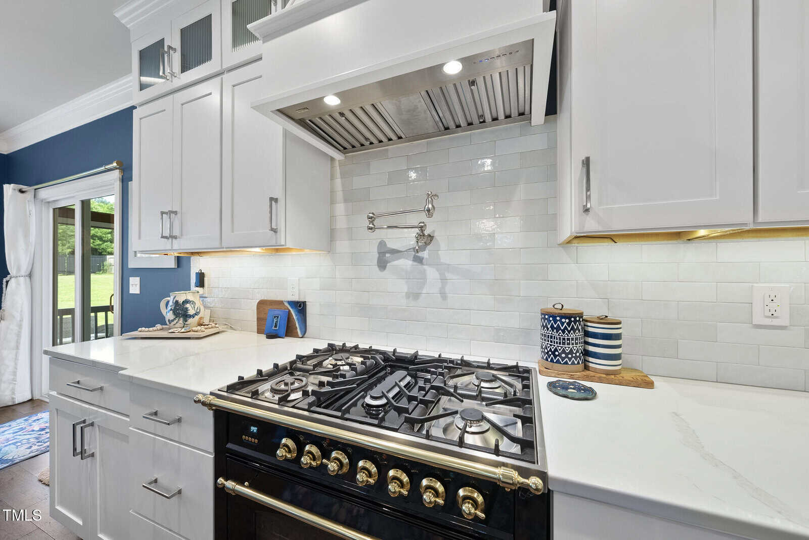 A modern kitchen features a black and gold gas stove with six burners, white cabinetry, white subway tile backsplash, and a stainless steel range hood. Blue and white canisters and a cutting board sit on the counter.