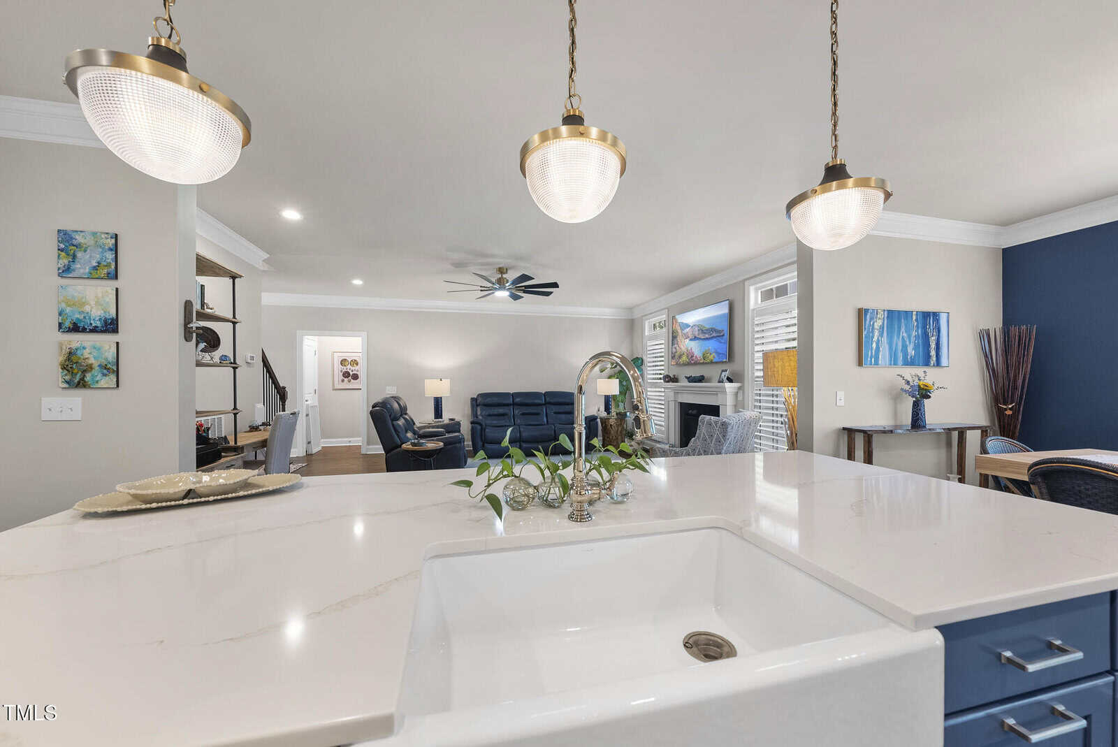 A modern open-concept living area with a white kitchen island and sink in the foreground, pendant lights overhead, blue and beige furniture, wall art, and a ceiling fan in the background.