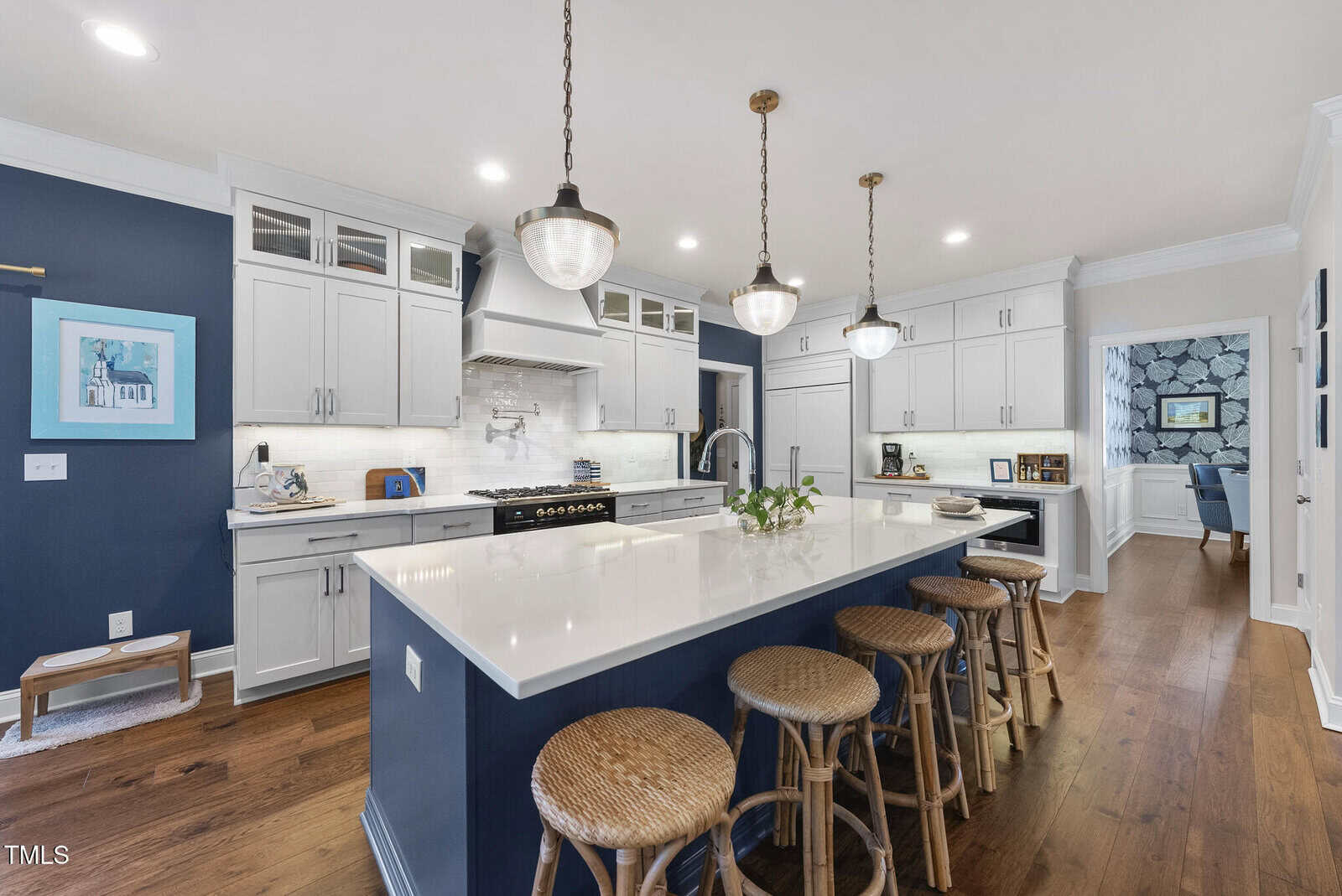 Modern kitchen with white cabinets, a large island with wicker stools, dark blue accent wall, hanging pendant lights, and wood flooring. A dining room with patterned wallpaper is visible through a doorway.