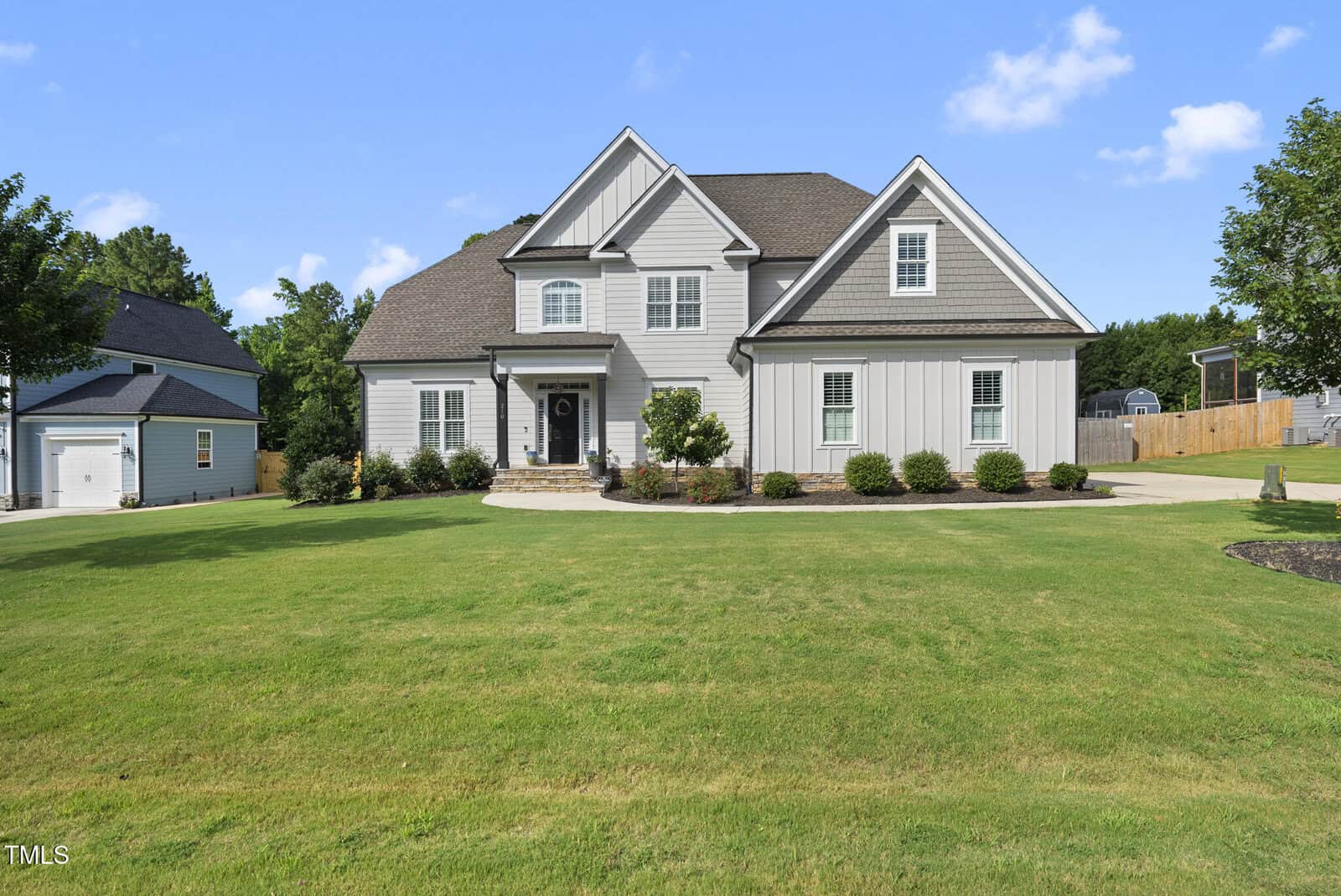 A large, two-story house with light gray siding, white trim, and a dark shingle roof stands behind a wide, neatly manicured lawn. Bushes line the front of the house and a driveway curves to the right.