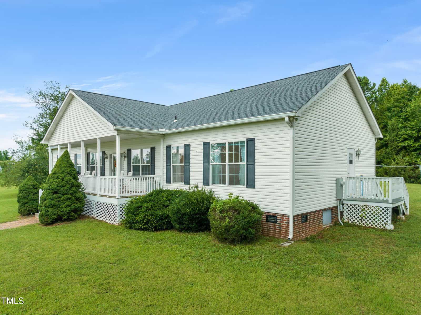 Single-story white house with dark shutters and a gray shingled roof, featuring a front porch and small deck, surrounded by green lawn and shrubs, on a clear day.