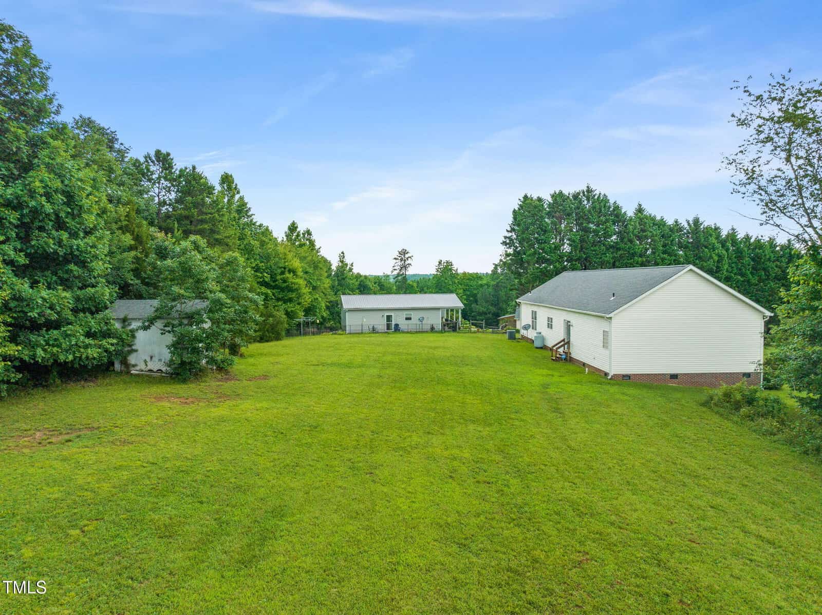 A large grassy yard with two white buildings, one in the foreground and one farther back, surrounded by trees under a blue sky.