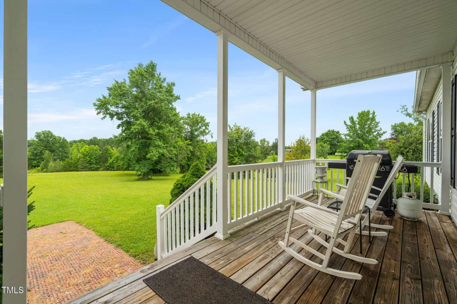 A covered wooden porch with two white rocking chairs and a grill overlooks a spacious green lawn with trees under a clear blue sky. White railings and steps lead down to a brick patio area.