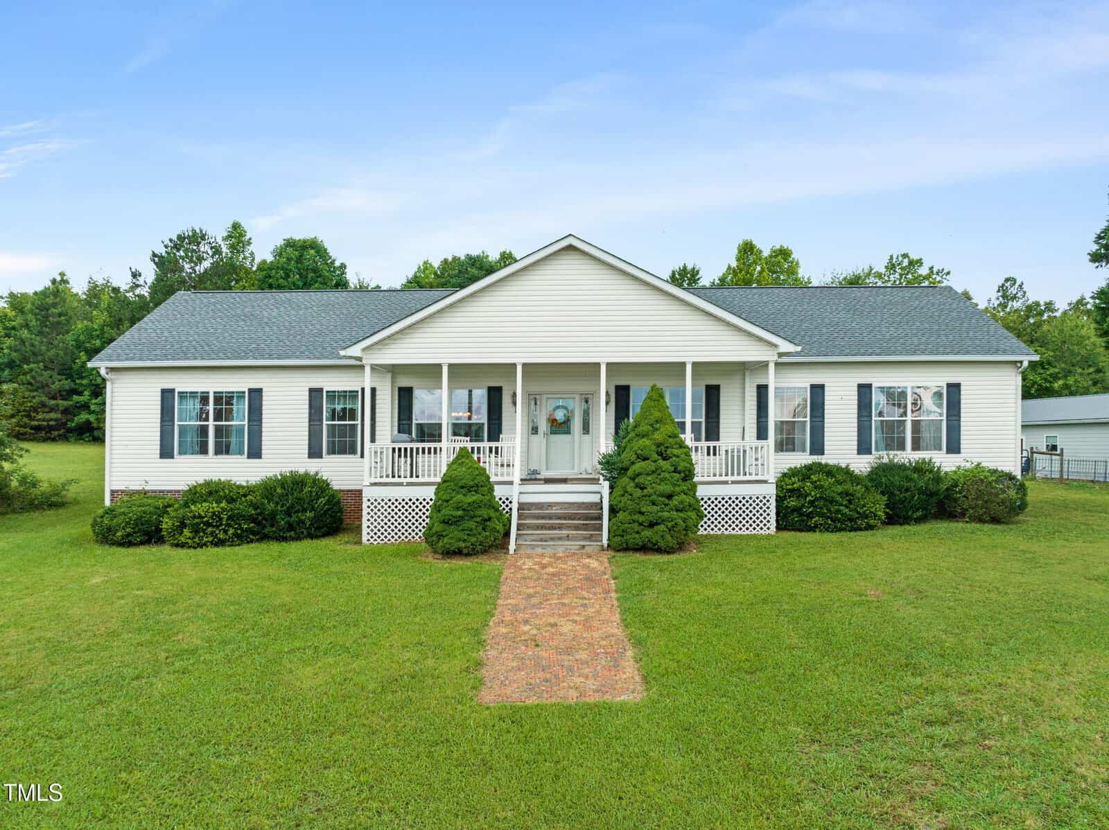 This charming single-story white house at 9633 Grassy Creek Road features a gray roof, front porch, and steps leading to a brick pathway. Green shrubs line the front lawn, with trees in the background under a clear Bullock blue sky.