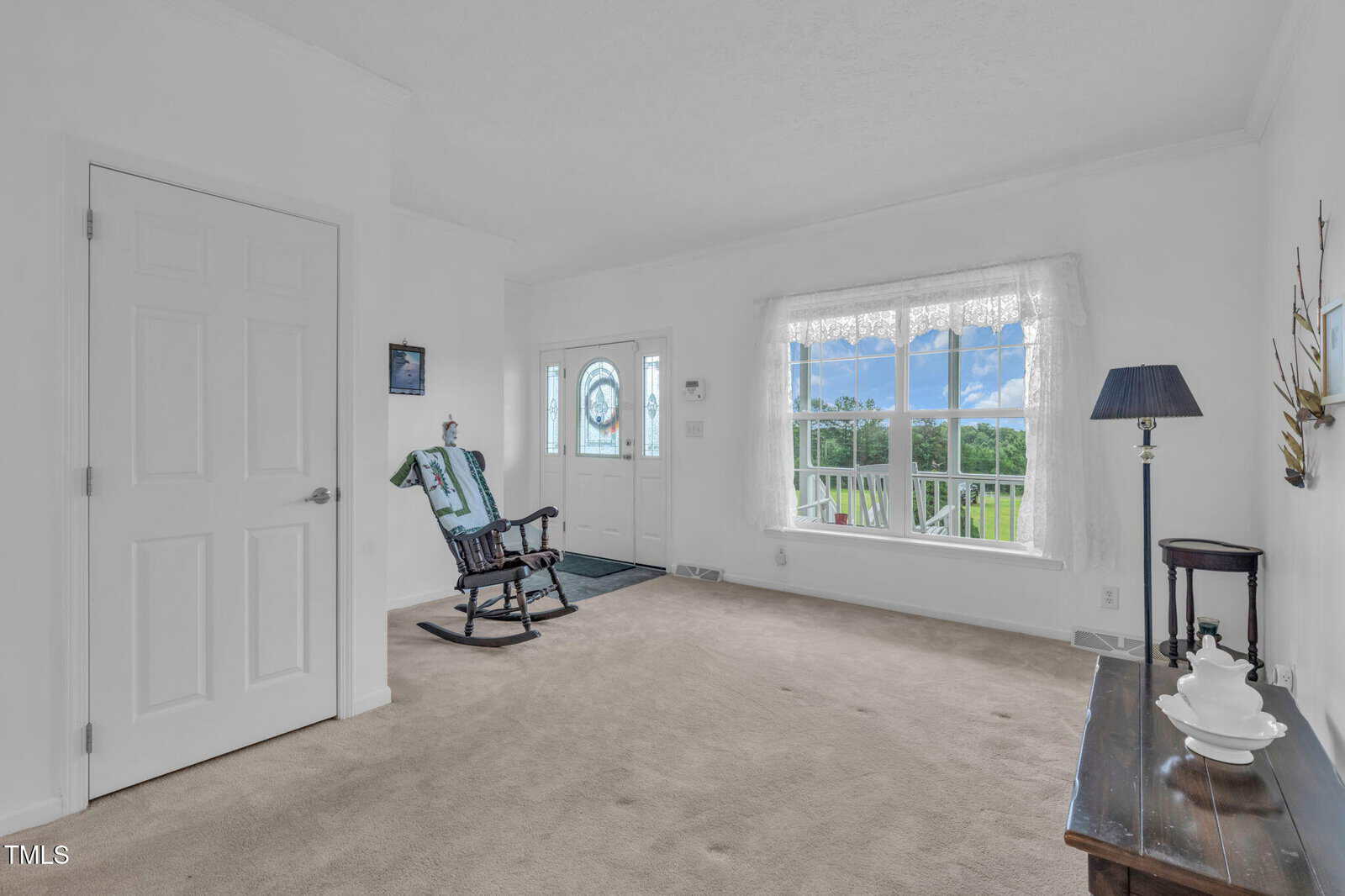 Bright living room with beige carpet, large window with sheer curtain, rocking chair near the door, small table with lamp, and outdoor view of trees and blue sky through the window.