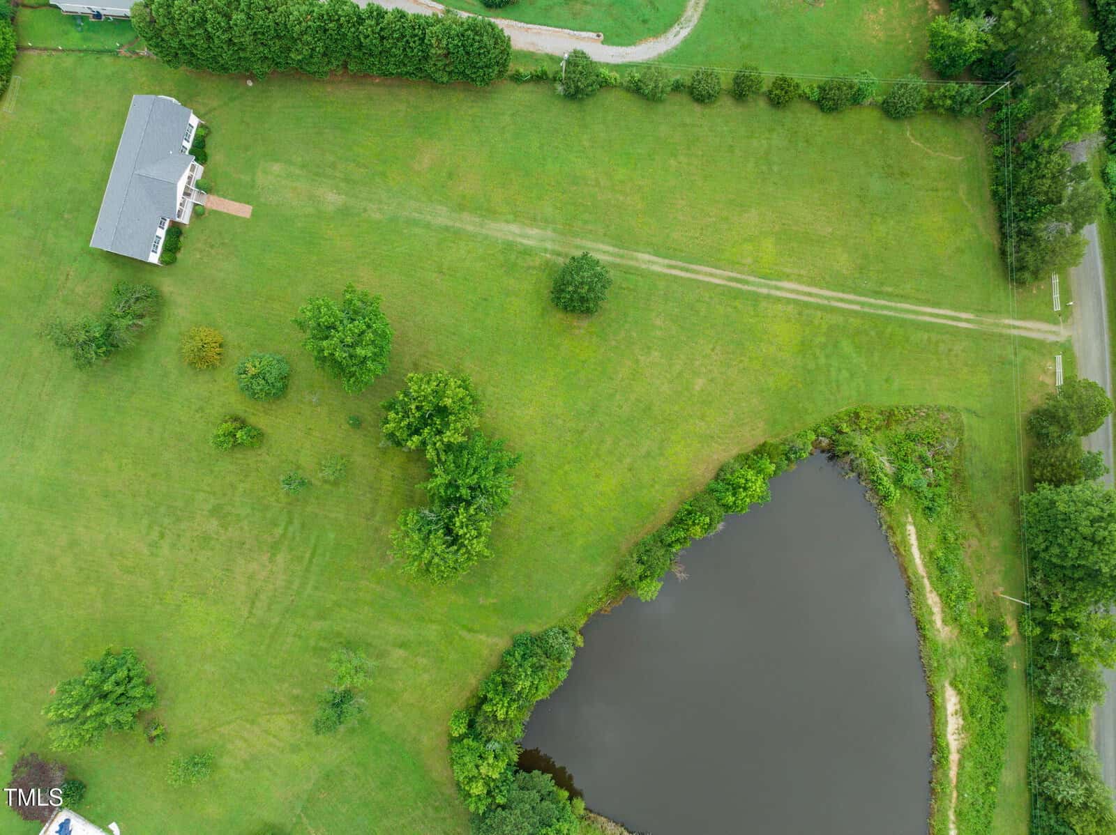 Aerial view of a grassy property with a house in the upper left, a pond in the lower right, scattered trees, and a winding driveway near the top.