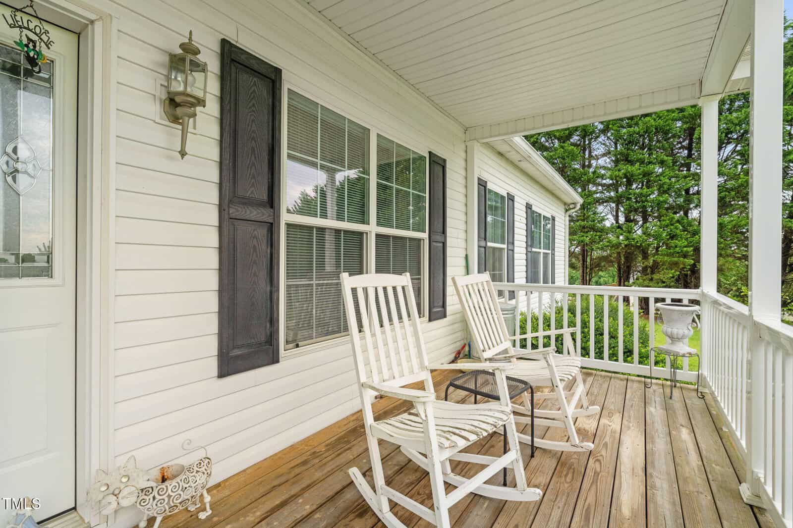 A white wooden front porch with two rocking chairs, a metal decorative pig, and a potted plant. The house has white siding, black shutters, and large windows. Trees and greenery are visible in the background.