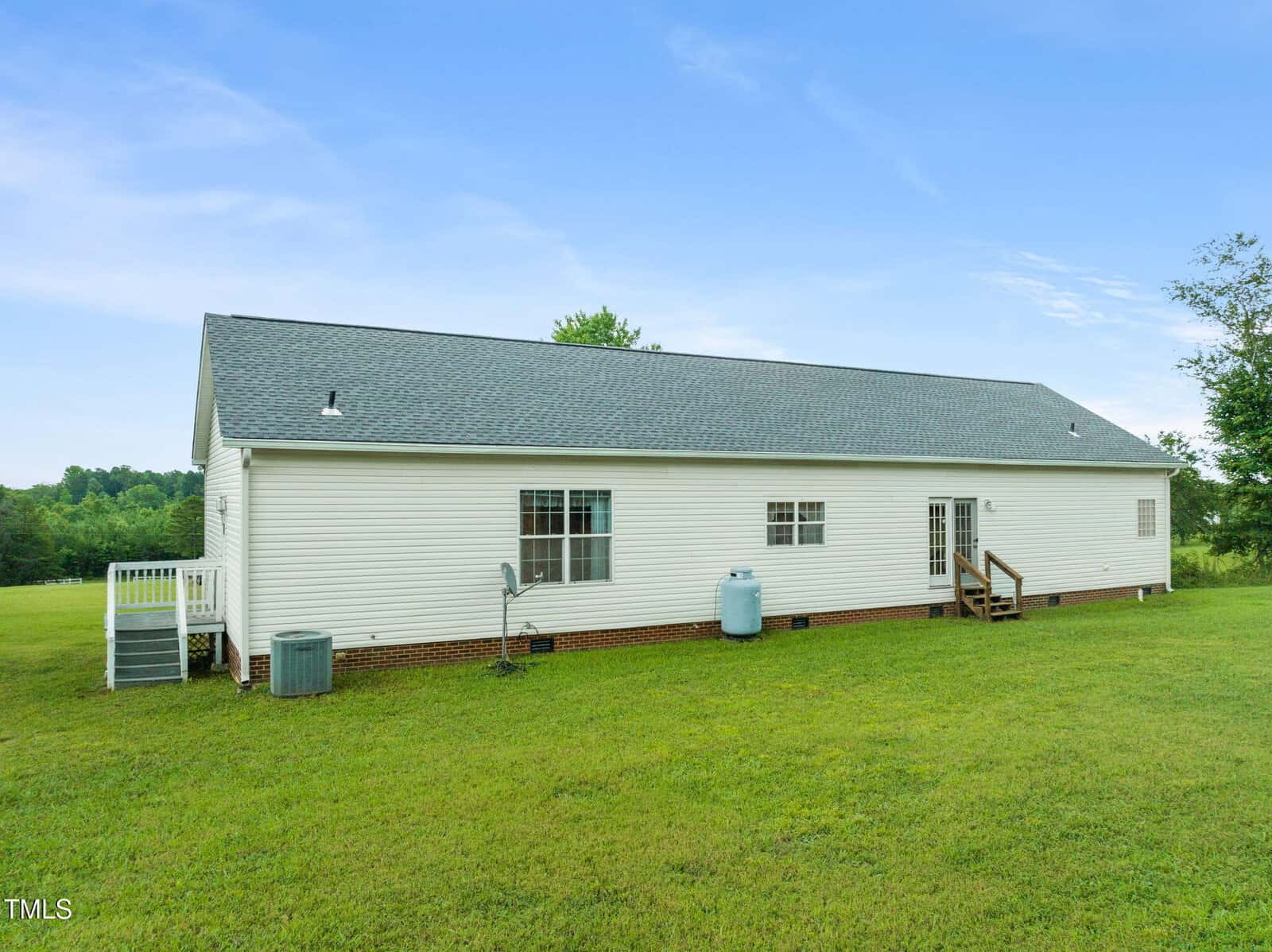 A single-story white house with gray shingles is shown from the back, featuring a grassy yard, a satellite dish, HVAC unit, propane tank, and two small porches with steps leading to doors. Trees and open land are in the background.