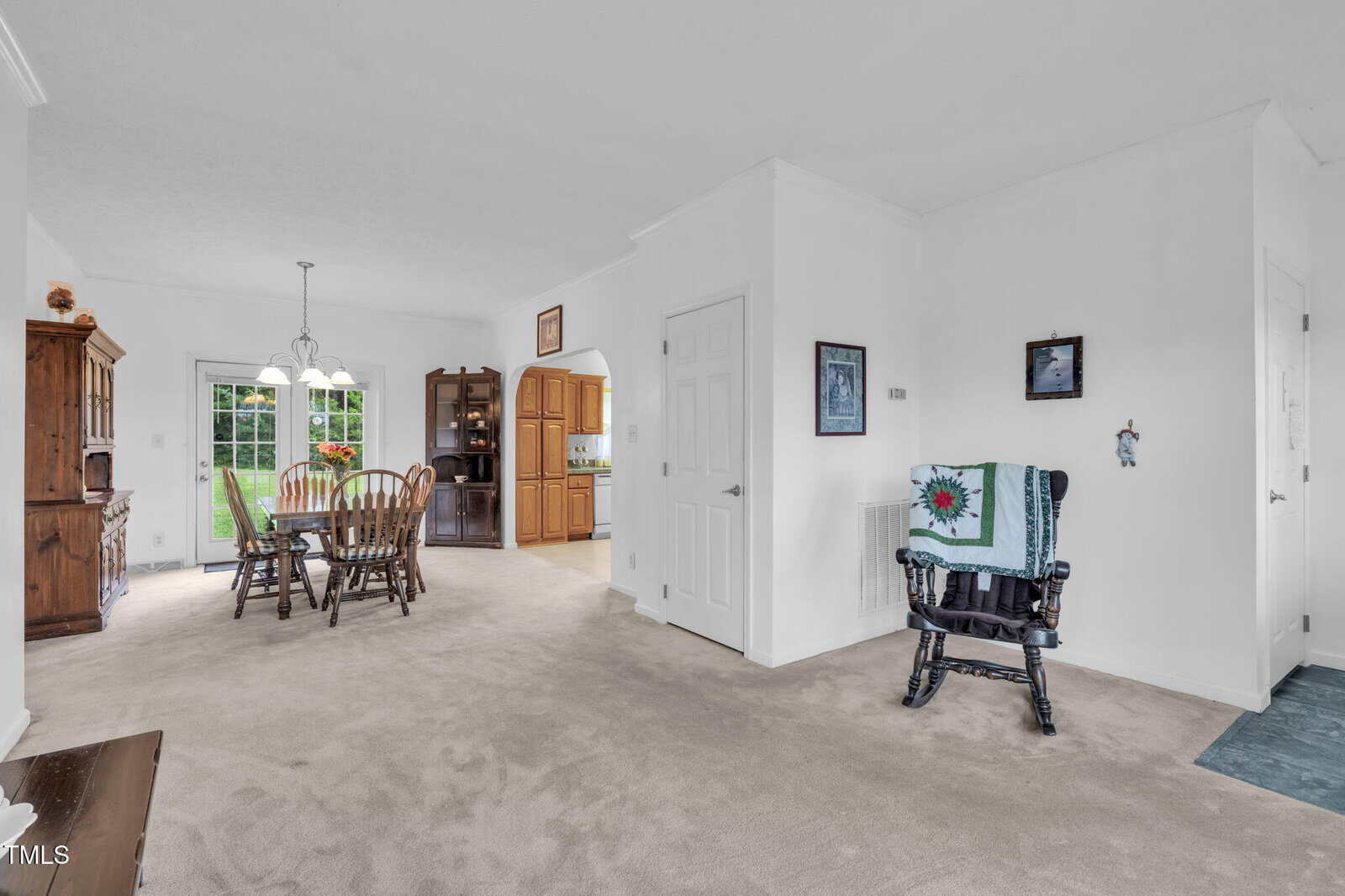 A dining room with a wooden table and chairs, a hutch, and a view into a kitchen. A black chair with a quilt draped over it sits by a white wall. Glass doors open to a green lawn outside.