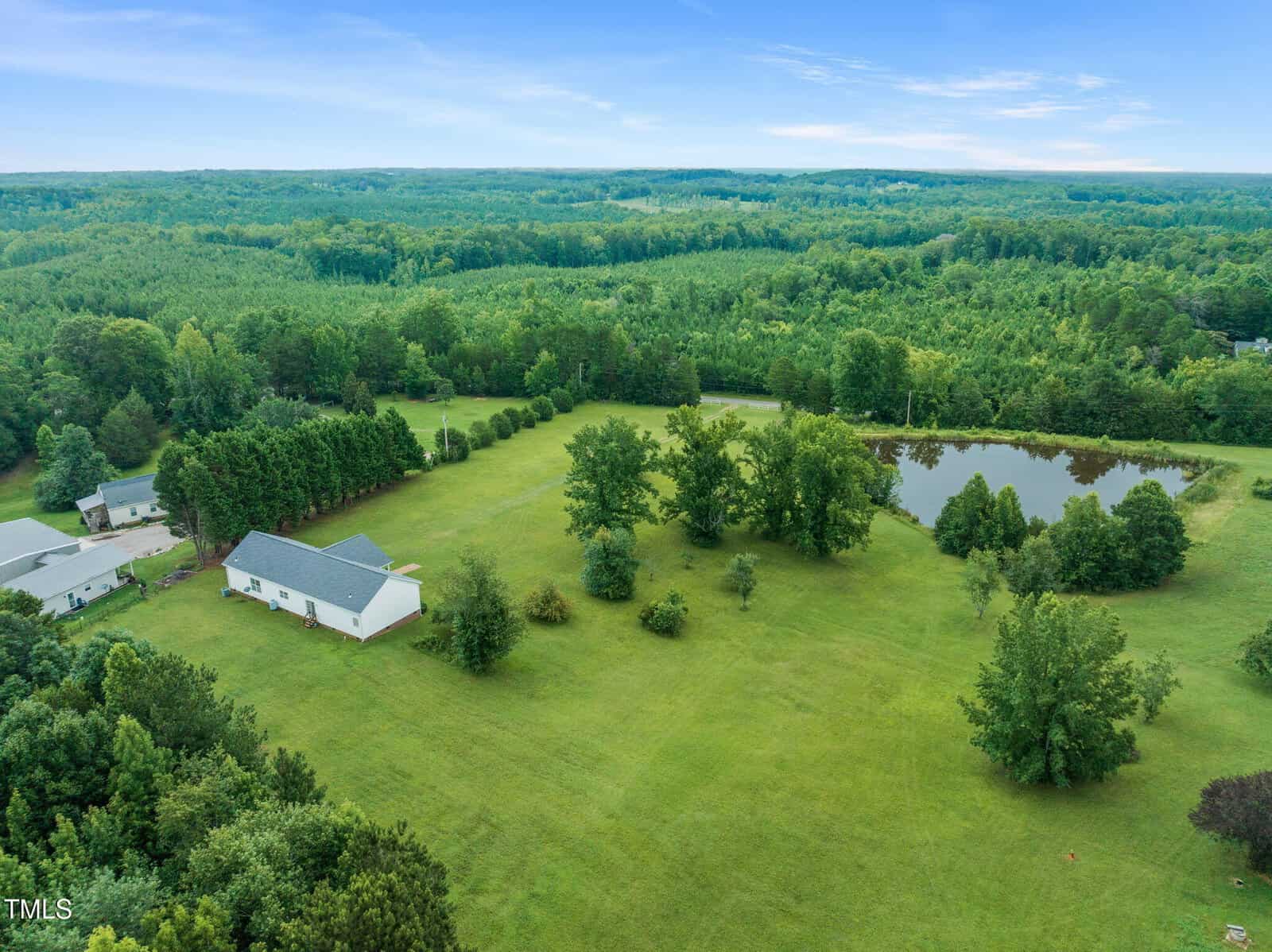 Aerial view of a large grassy property with scattered trees, a white house with a gray roof, a nearby pond, and dense forest in the background under a blue sky with light clouds.