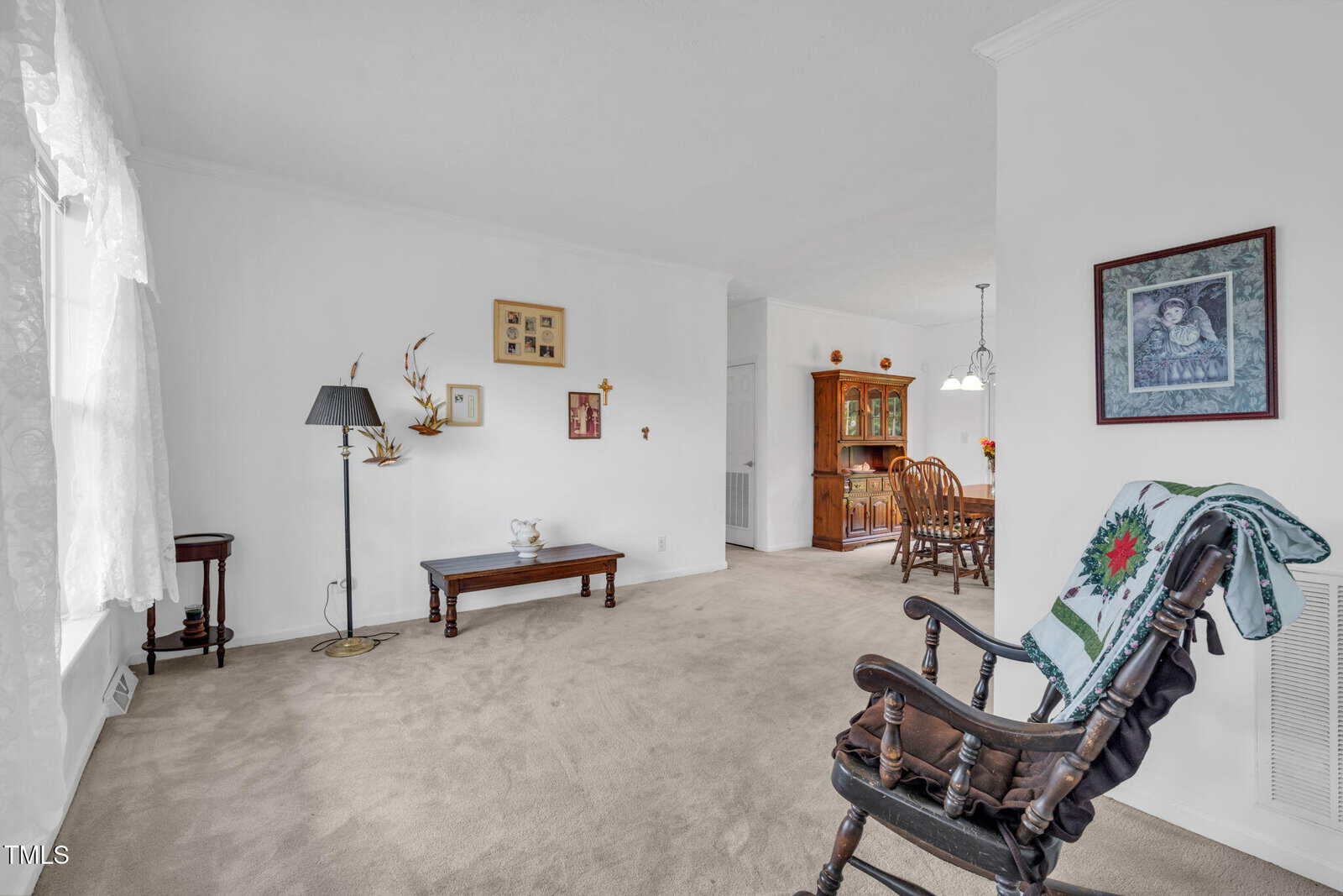 A living room with beige carpet, a wooden rocking chair with a quilt, a bench, a floor lamp, and wall art. In the background is a dining area with a wooden table, chairs, and a hutch.