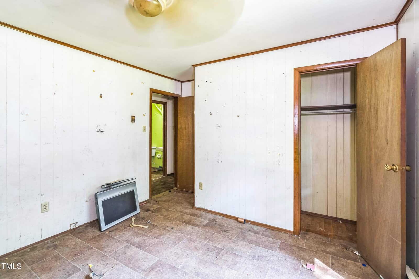 A mostly empty room with white paneled walls, tan ceiling, and brown trim. The floor is bare with some debris. An old TV lies on the floor, the closet door is open, and a second door leads to another room.