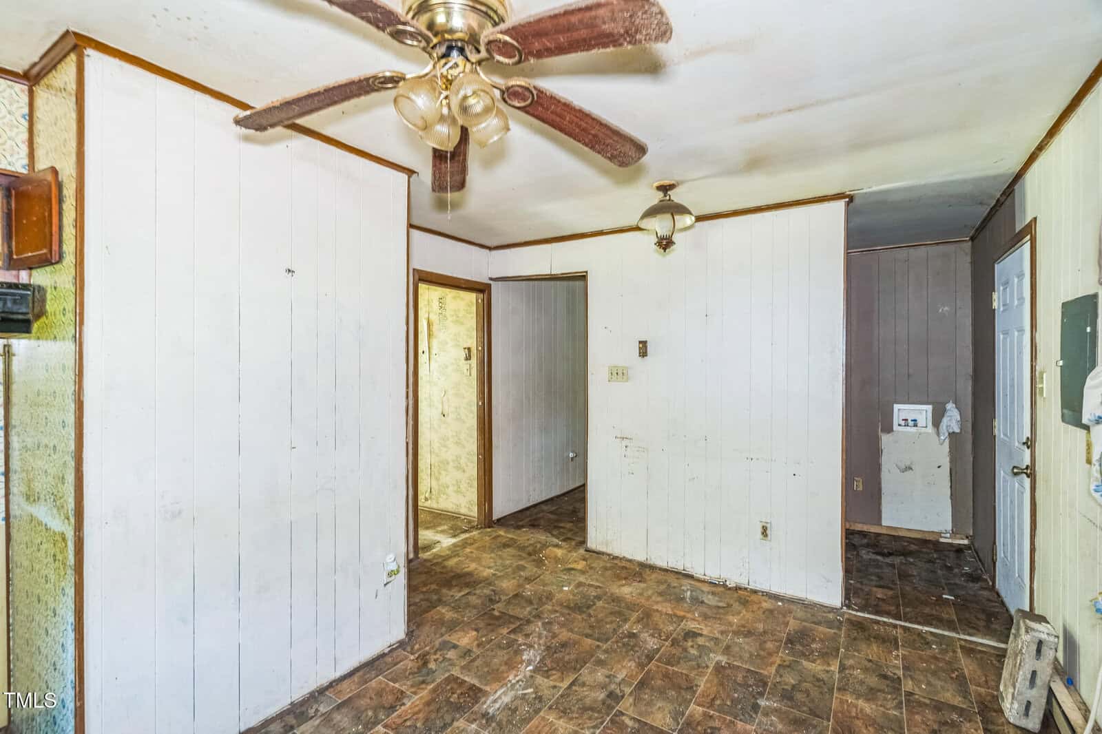 A room with white wood-paneled walls, a ceiling fan with four blades, brown tiled floor, a hanging light fixture, visible dirt and wear on surfaces, and doorways leading to other areas.