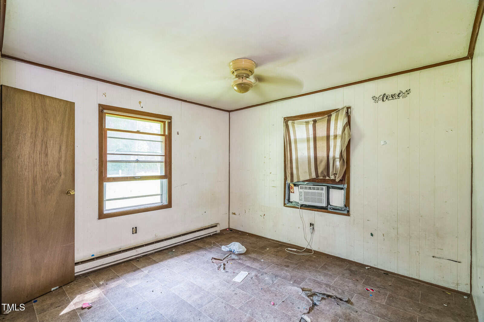 Empty, worn-out room with dirty walls, a ceiling fan, an air conditioner in the window with a striped curtain, scattered debris on the floor, and an open wooden door. Natural light enters through two windows.