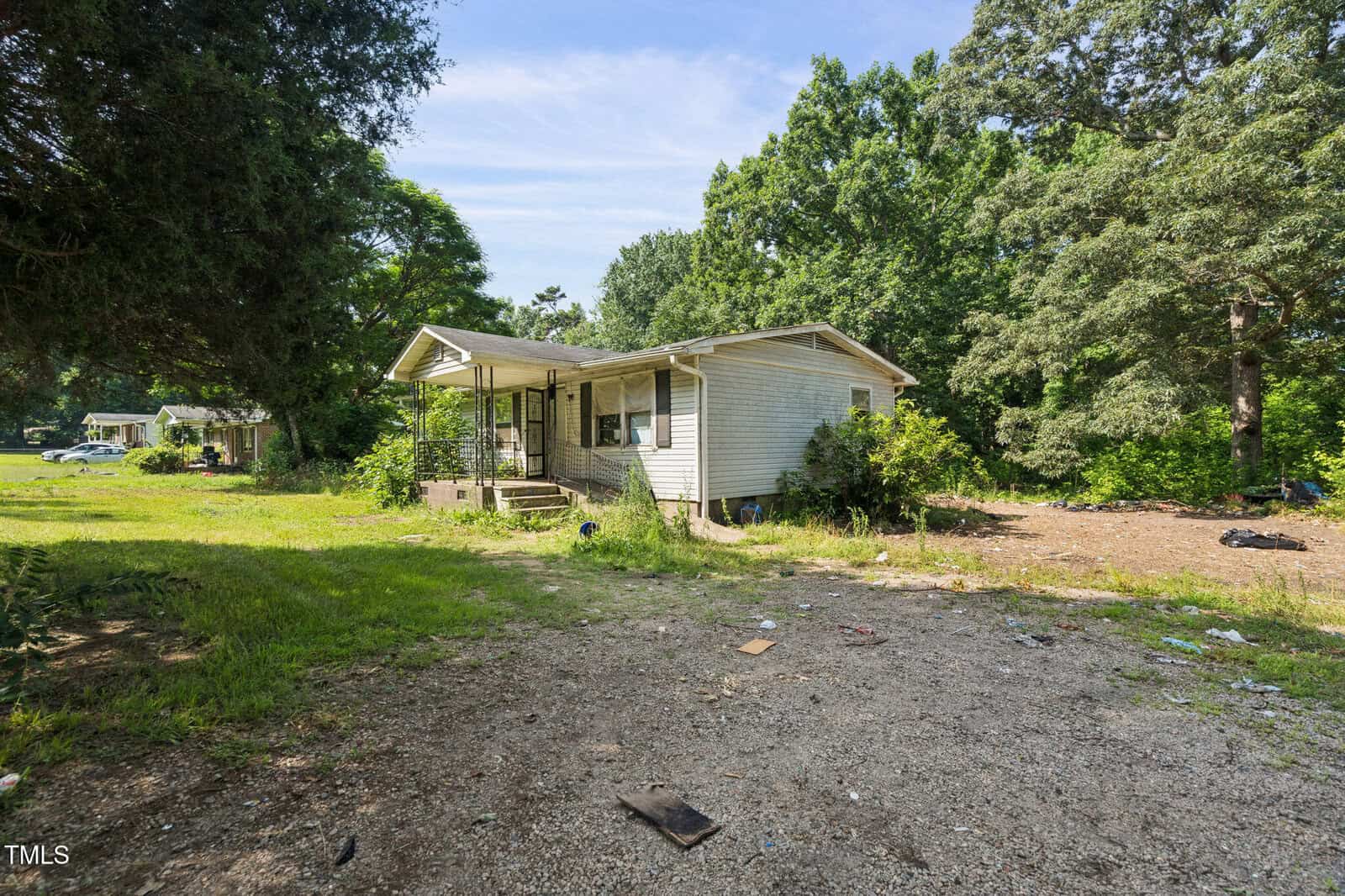 A small, single-story house with a covered porch sits on a grassy lot surrounded by trees. The yard is scattered with debris, and another house is visible in the distance on the left.