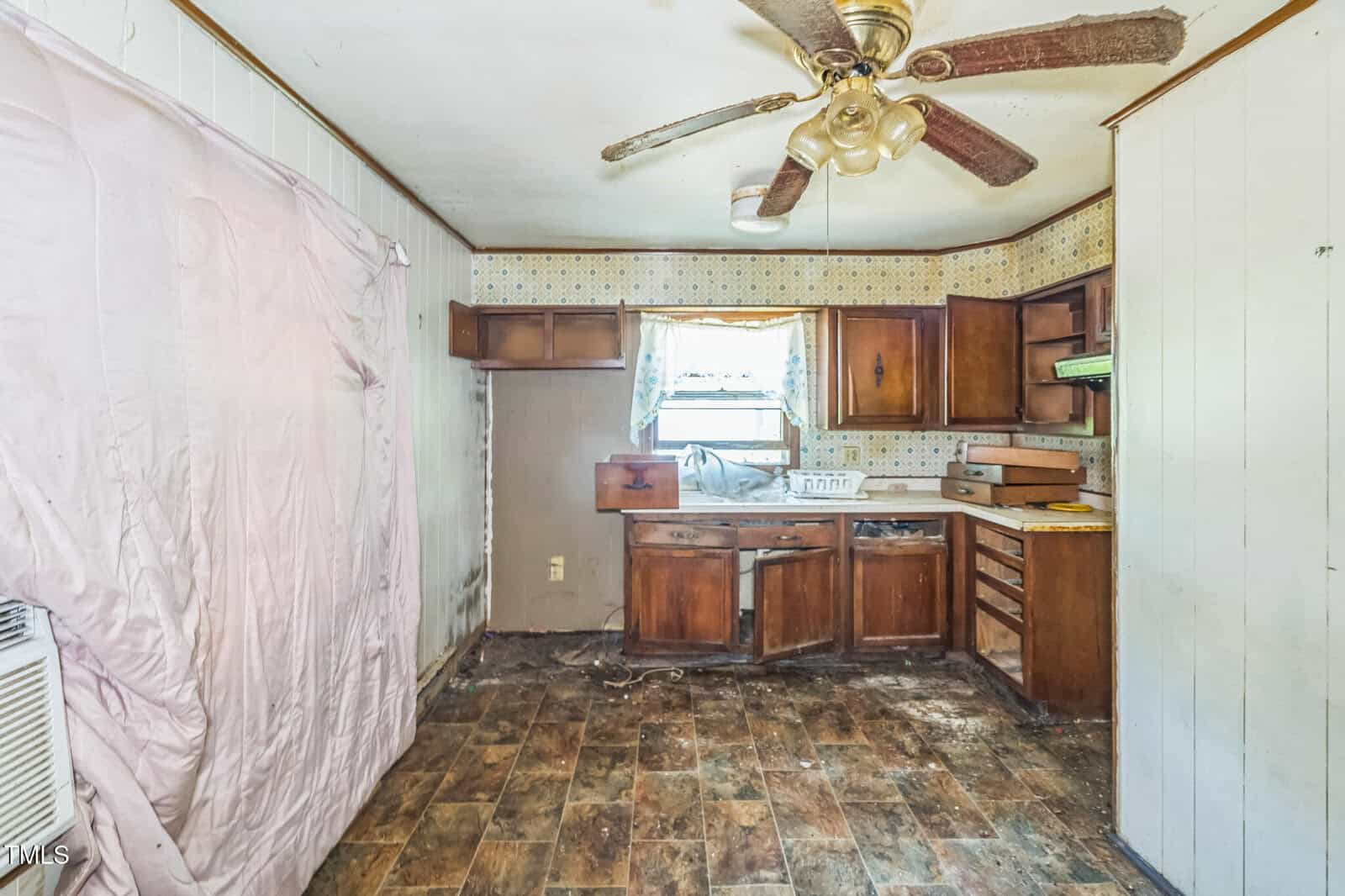 A worn kitchen with broken cabinets, dirty floors, a ceiling fan with missing blades, and stained walls. A pink sheet hangs on the left side. Natural light enters through a window above the sink.