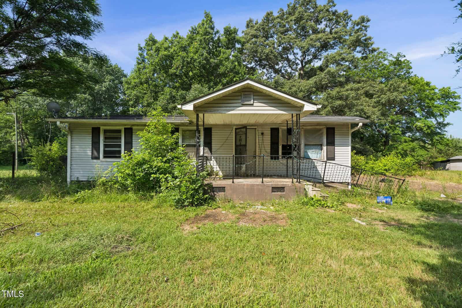 Nestled at 1141 Morgan Road in Henderson, this small, single-story white house features a covered front porch with metal railings, surrounded by overgrown grass and towering trees under a clear blue sky.