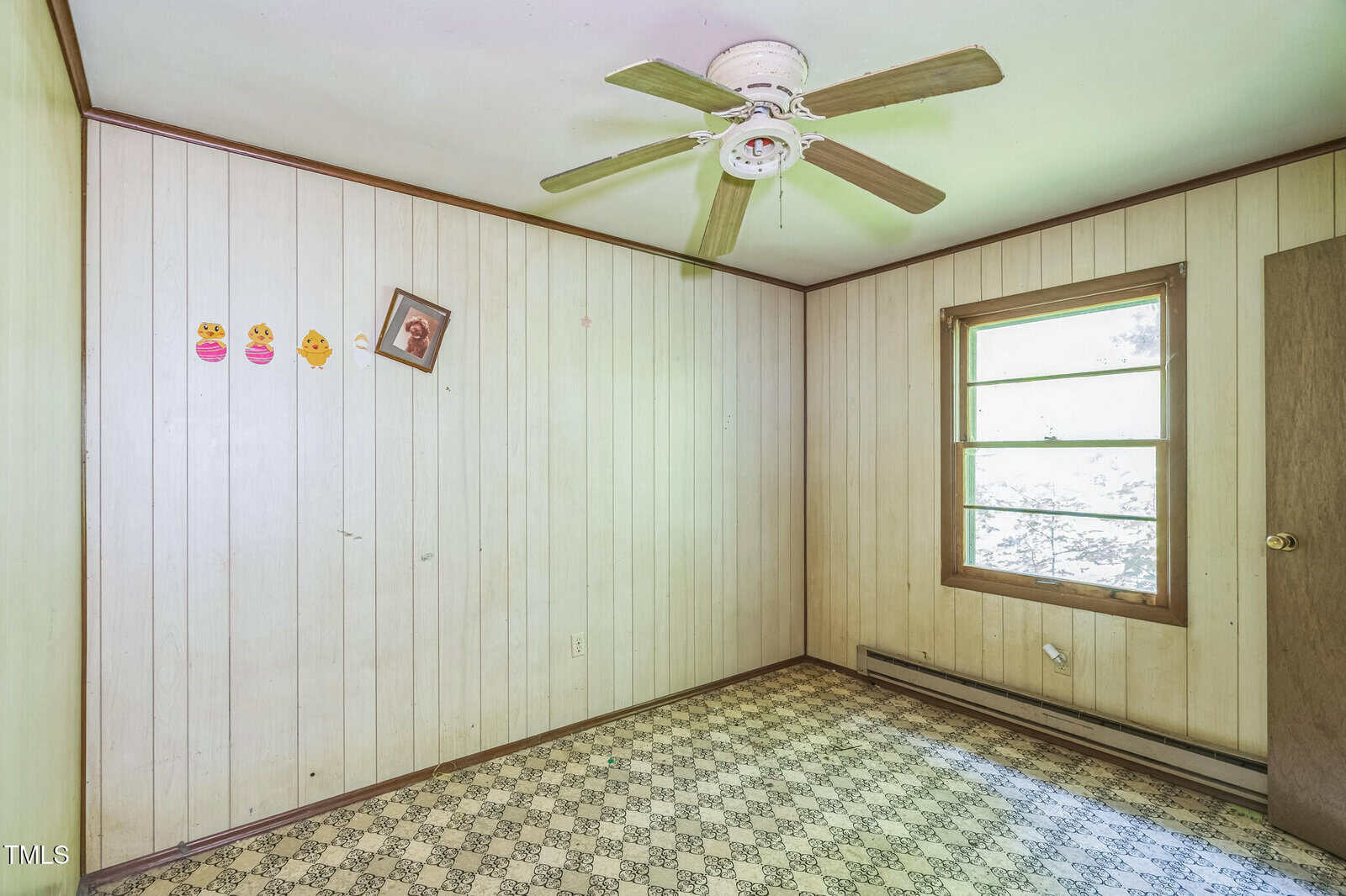 A small, empty room with wood panel walls, a patterned linoleum floor, a ceiling fan, a window, a door, three emoji stickers, and a crooked framed picture on the wall.