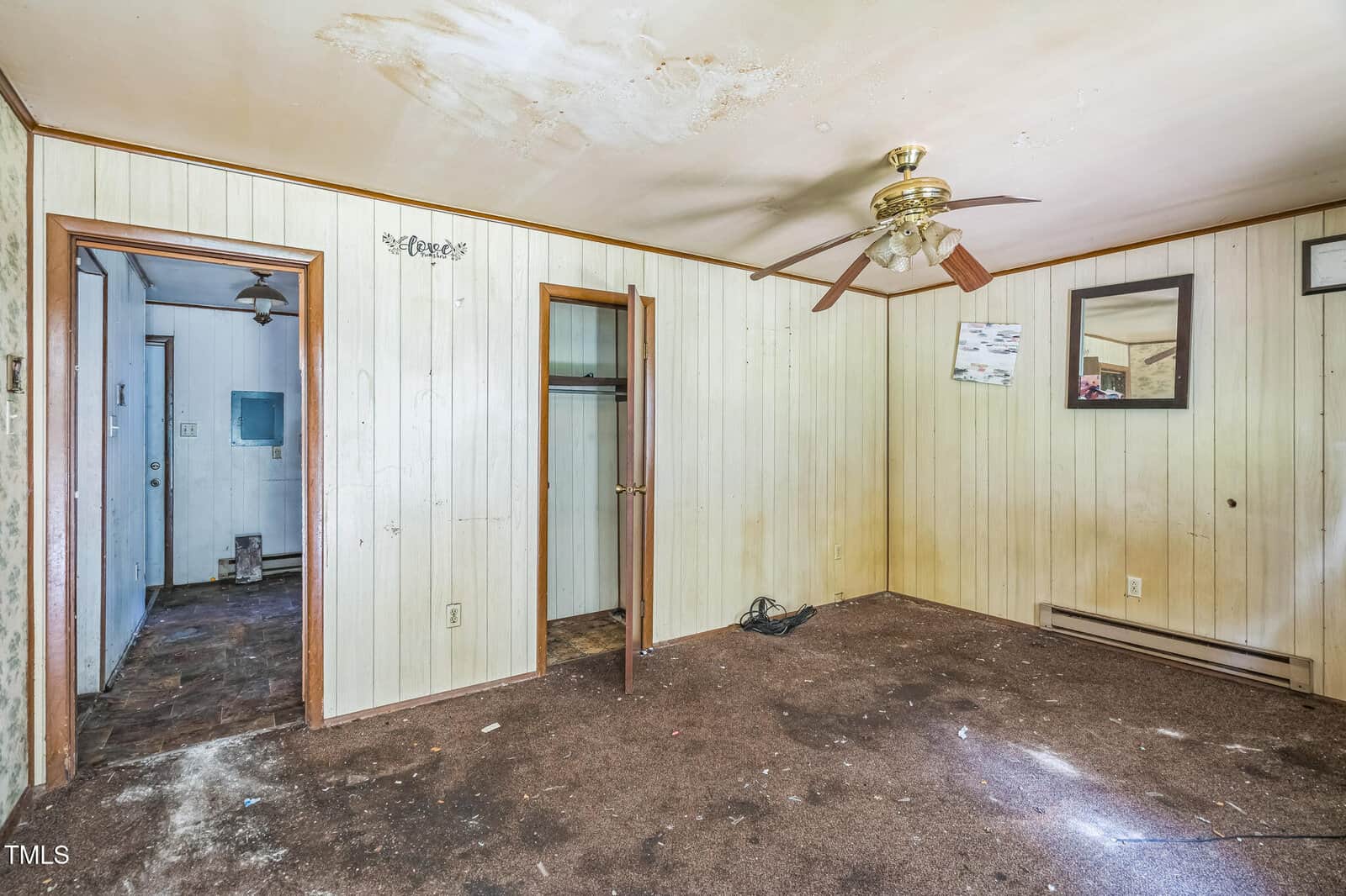 Empty, worn room with stained carpet, yellowed wood-paneled walls, ceiling fan, open closet, and another doorway leading to a messy adjoining space. A few wall hangings and visible ceiling damage.