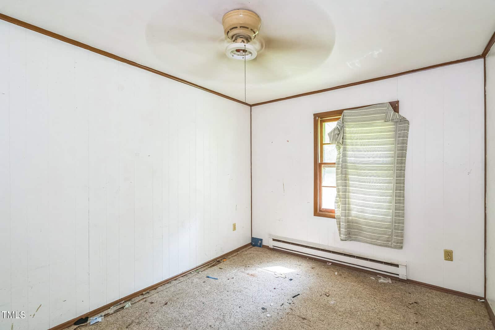 Empty room with white paneled walls, beige carpet, a ceiling fan, a single window with a wrinkled curtain, and some scattered debris on the floor.