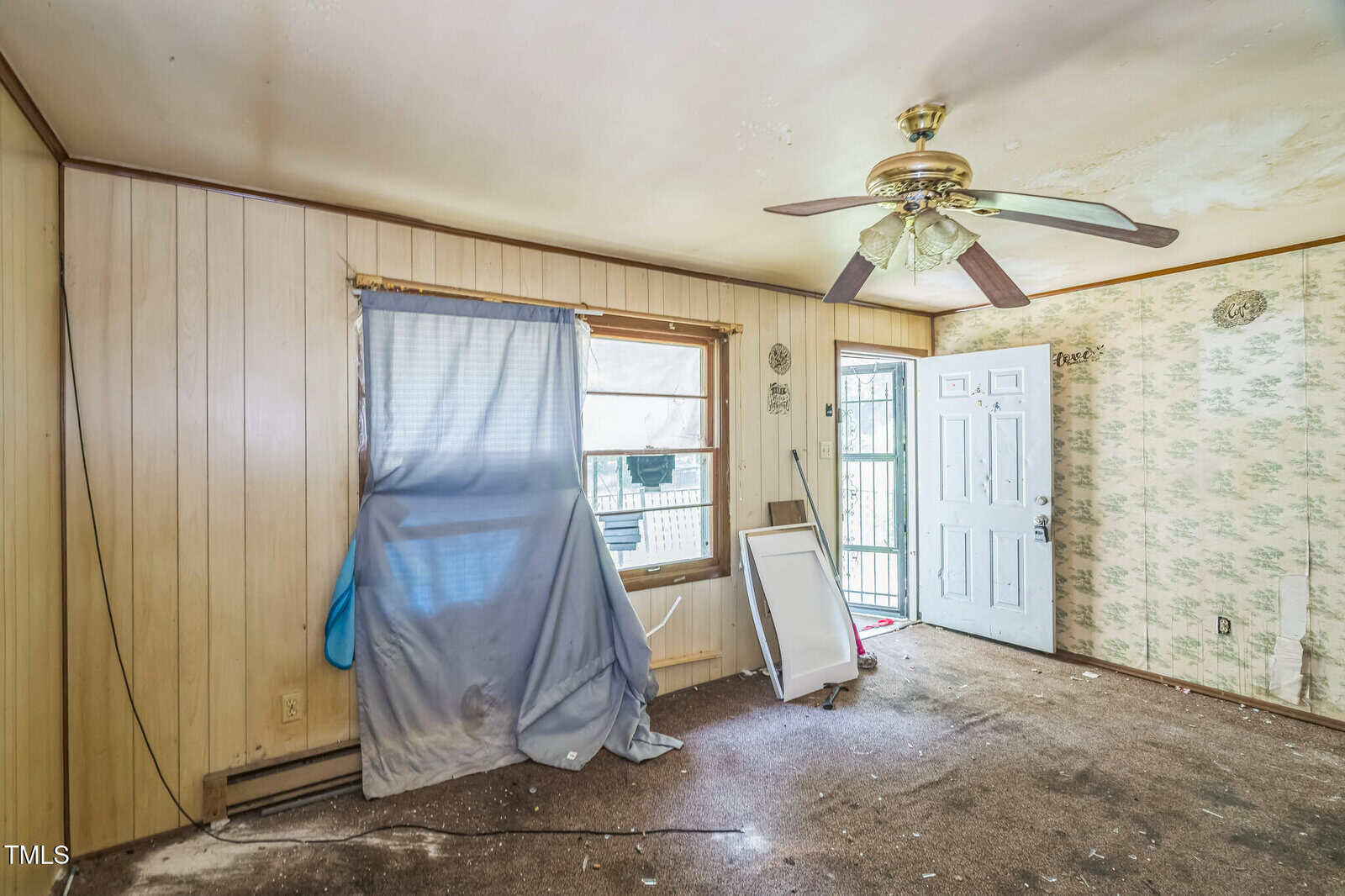 A small, cluttered room with wood panel walls, a ceiling fan, a window partially covered by a curtain and debris, a damaged screen, an open door, and stained carpeting. Walls show peeling wallpaper and dirt.