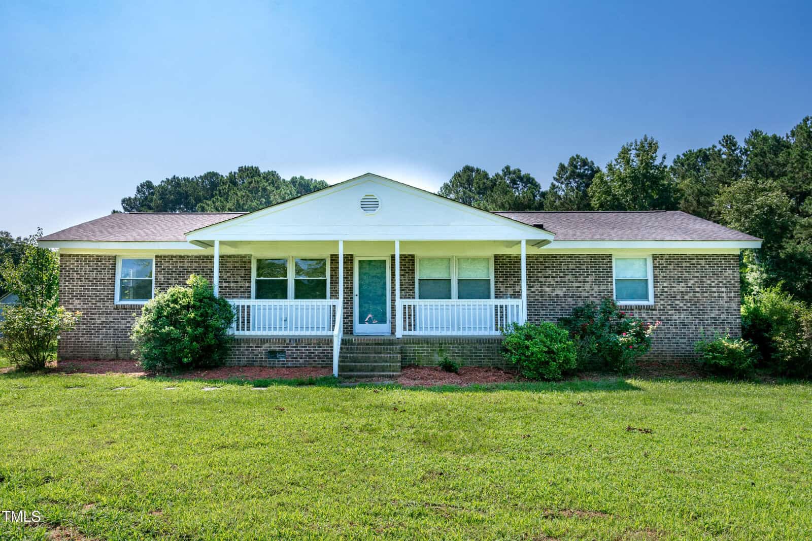 Single-story brick house with white trim and a covered front porch, surrounded by green shrubs and grass, set against a backdrop of trees under a clear blue sky.